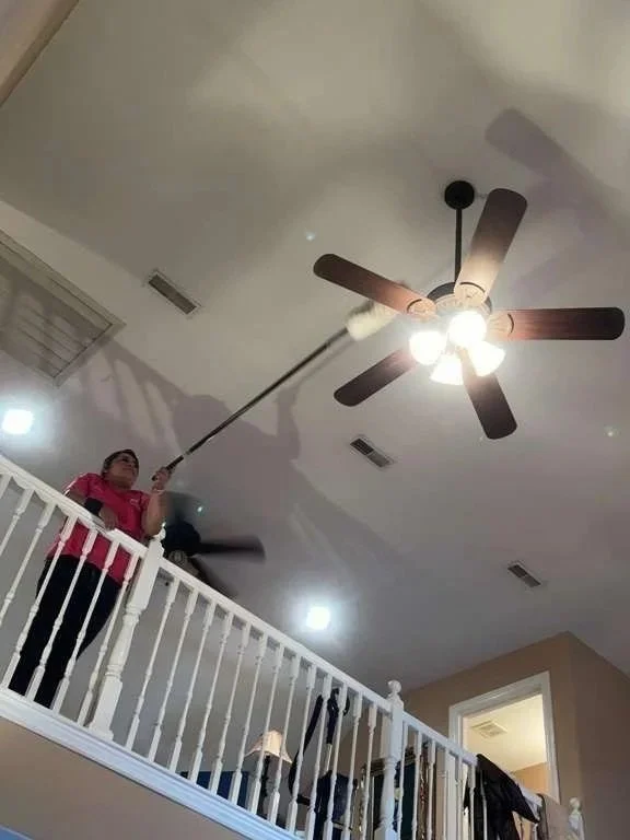 A woman cleaning a ceiling fan with a long duster while standing on a balcony railing inside a home.
