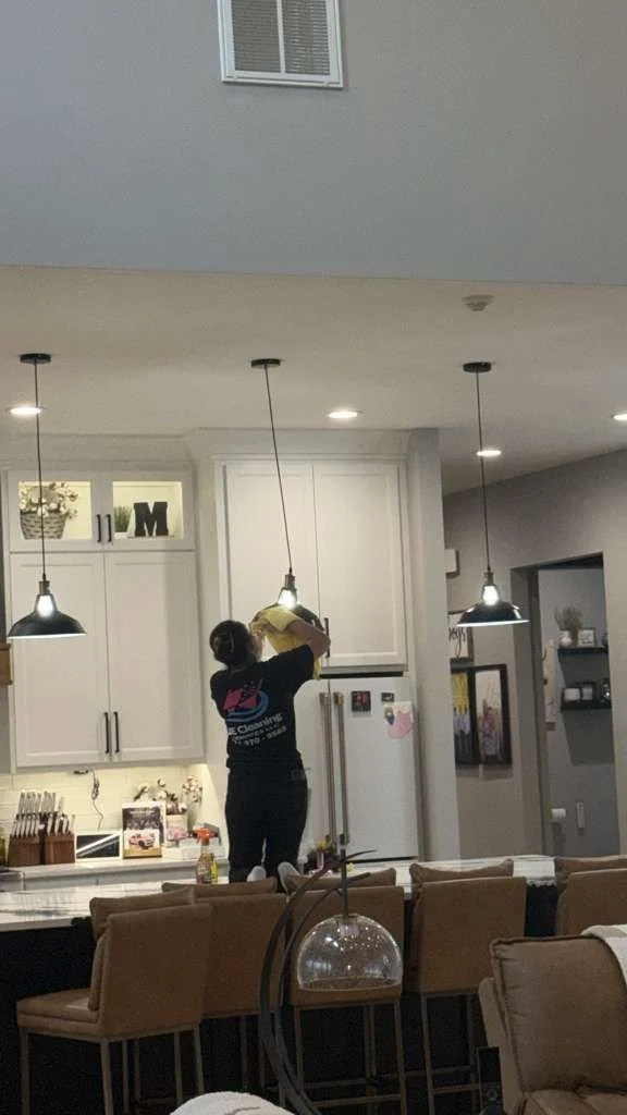 A woman cleaning the kitchen ceiling while standing on a chair in a modern kitchen with white cabinets, a white refrigerator, and a large island with beige chairs.