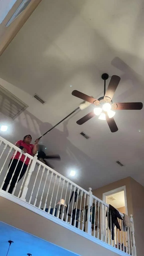 A woman in a pink shirt standing on a balcony cleaning the ceiling fan with a long-handled duster.