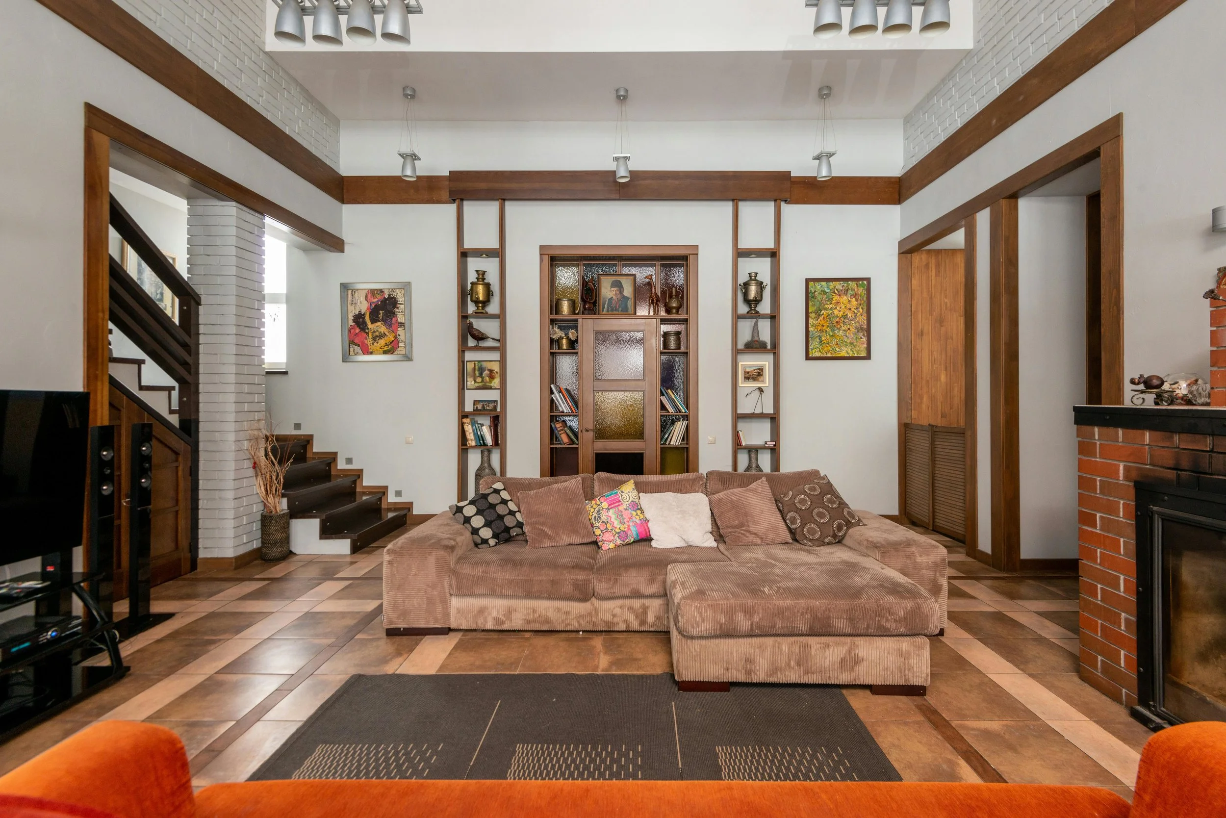 A cozy living room with a large brown sectional sofa adorned with colorful pillows. Behind the sofa, there is a built-in wooden wall unit with shelves displaying decorative items, books, and framed pictures. The room features white walls with wooden trim, stained glass panels, and artwork. To the right, a brick fireplace is partially visible. The floor has a geometric patterned wood design, and there are modern ceiling lights.