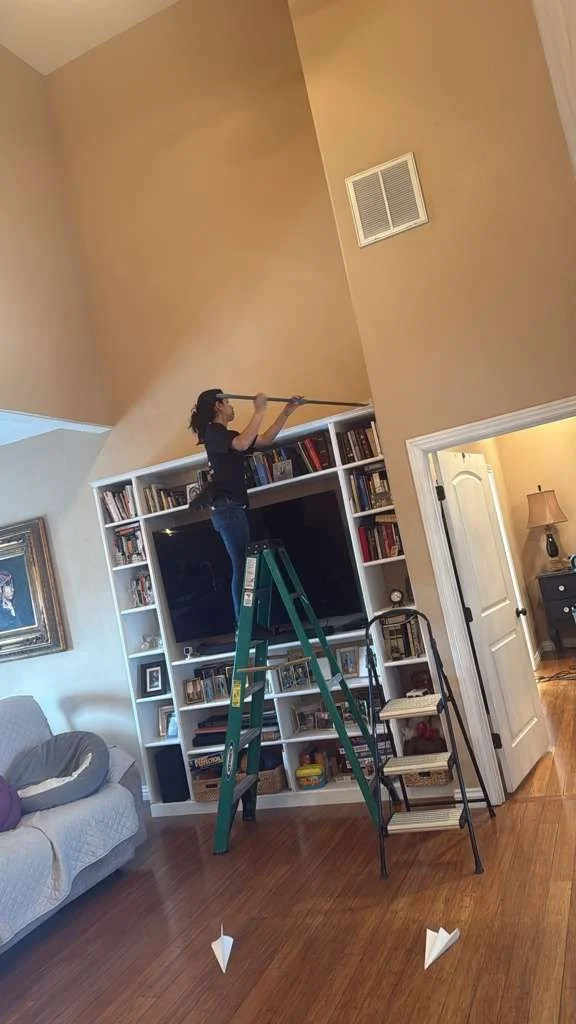 Woman standing on a step stool cleaning or reaching for a high shelf with a long-handled tool in a living room.