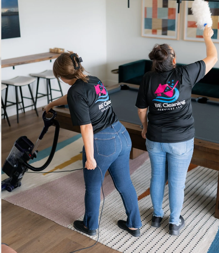 Two women wearing black t-shirts with a cleaning logo, one vacuuming and the other wiping a pool table, in a modern living room.