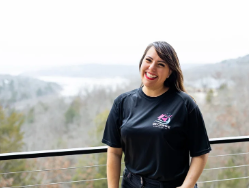 A smiling woman with long dark hair standing outdoors on a balcony with a scenic view of trees and a body of water in the background.