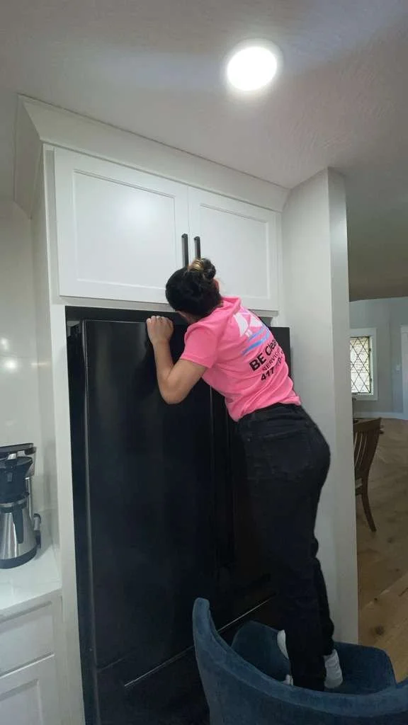 A person standing on a chair, reaching into a black refrigerator in a kitchen, wearing a pink shirt and black pants.