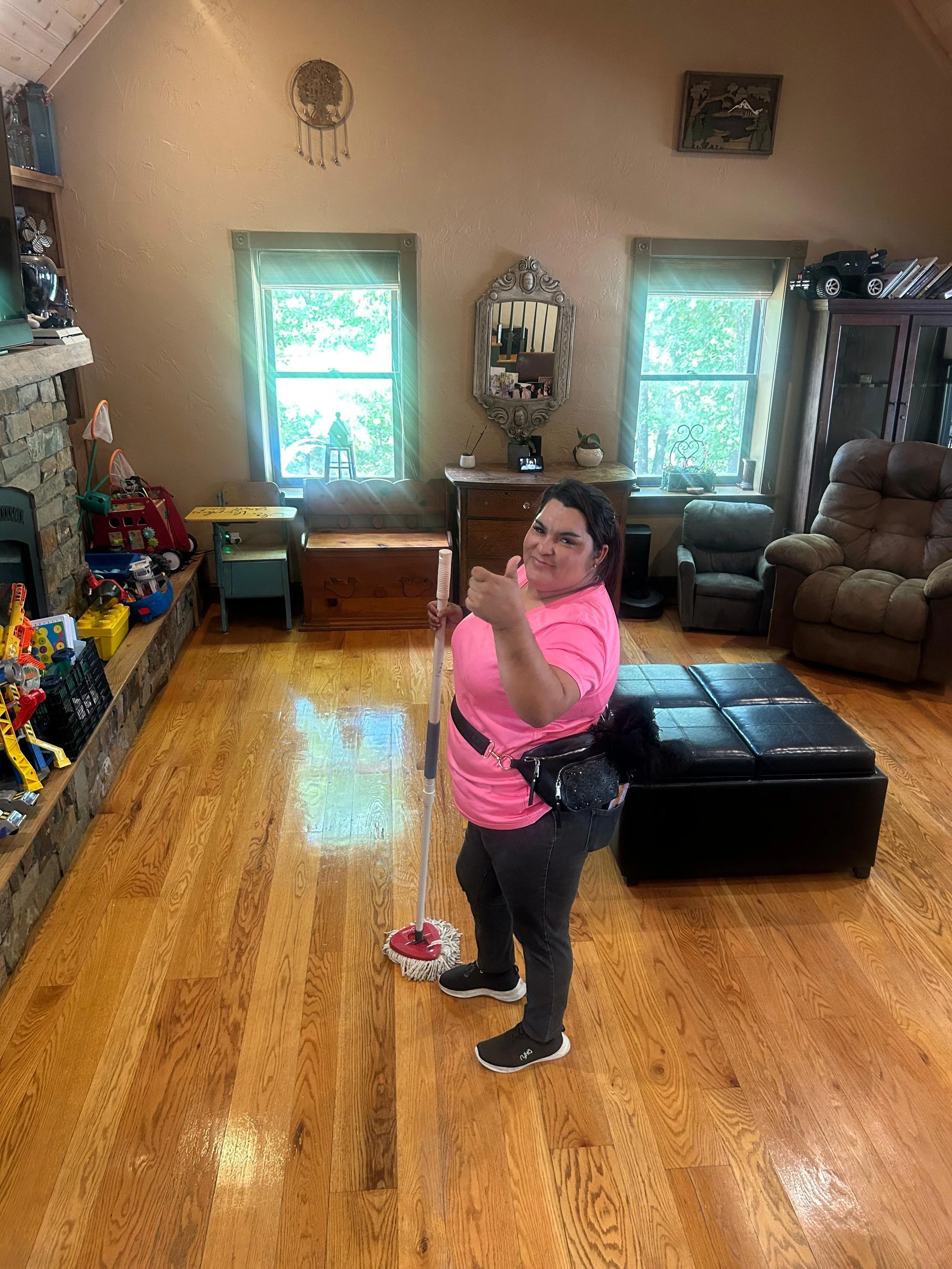 A woman in a pink shirt and black pants is standing on a hardwood floor, holding a mop with a red base and white mop head, giving a thumbs up. The room has two windows with greenish blinds, a stone fireplace, and various furniture and toys. She appea