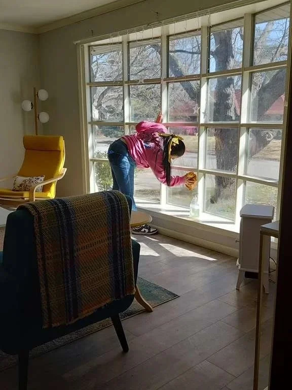 A woman cleaning a large window in a bright living room, with a yellow chair and an armchair with a plaid blanket.
