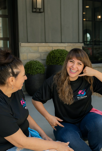 Two women laughing and talking outdoors, wearing matching black T-shirts with a pink and blue logo.