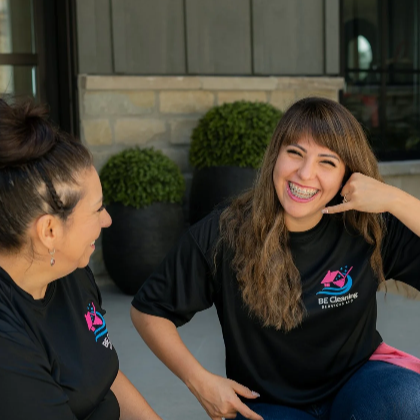 Two women sitting outdoors, engaged in conversation, smiling, wearing black t-shirts with a logo, with a building and plants in the background.