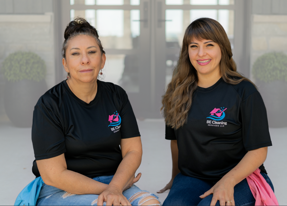Two women sitting indoors, wearing matching black T-shirts with the 'BE Cleaning Services LLC' logo, smiling and looking at the camera.