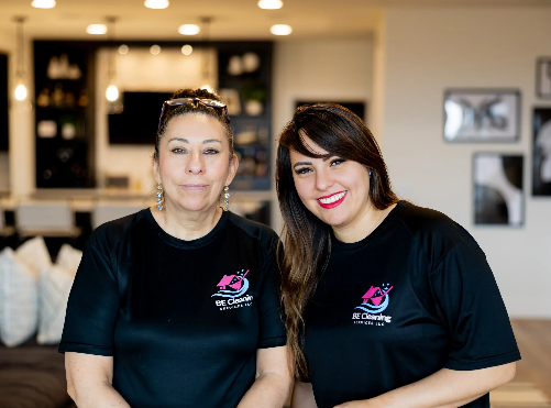 Two women in black T-shirts with a logo, smiling inside a modern, well-lit room with framed pictures on the wall.