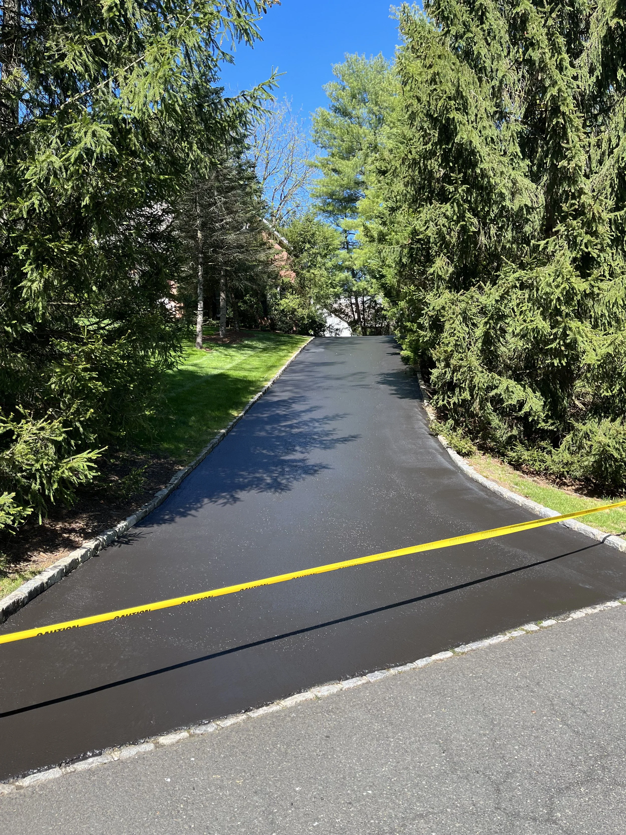 A newly paved asphalt road with a yellow 'Caution' tape across it, surrounded by green trees and grass, with a clear blue sky overhead.