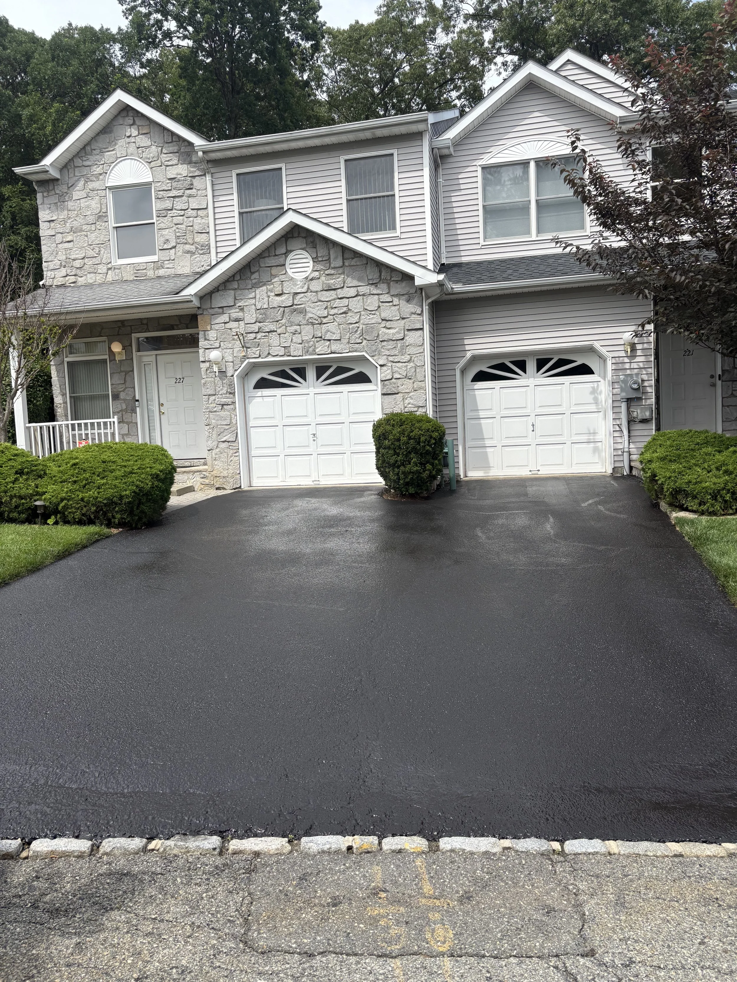 Two-story house with stone and siding exterior, two garage doors, shrubs in front, trees in the background, and freshly paved driveway.
