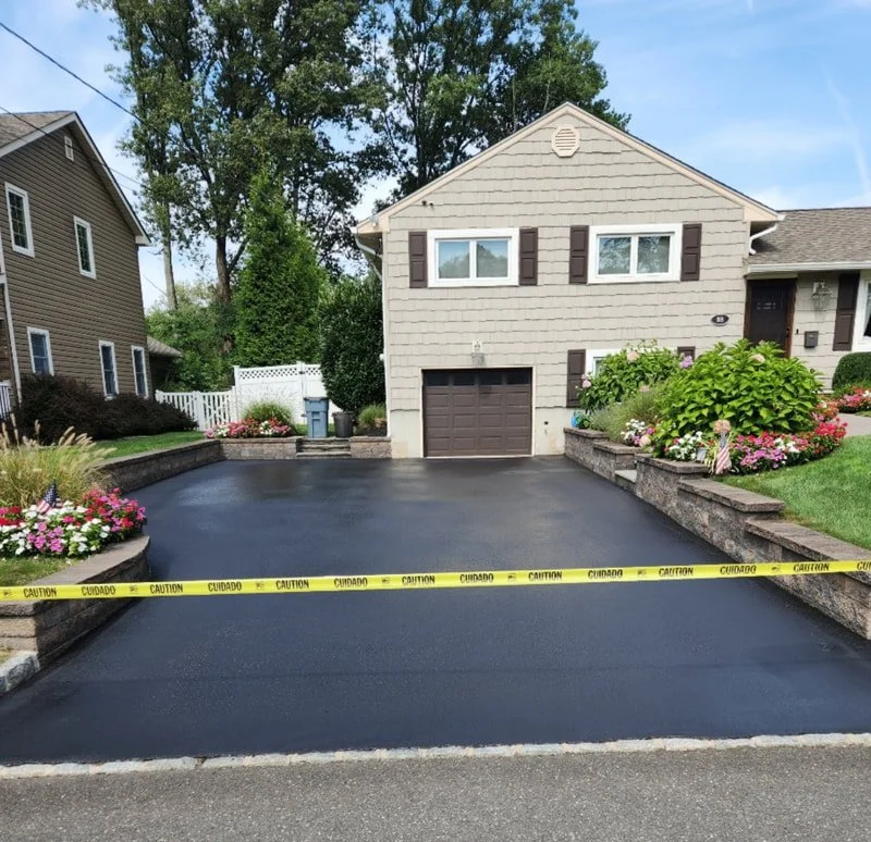 Freshly paved black asphalt driveway in front of a beige house with brown shutters, surrounded by colorful flowers and landscaped garden beds, with caution tape across the front.