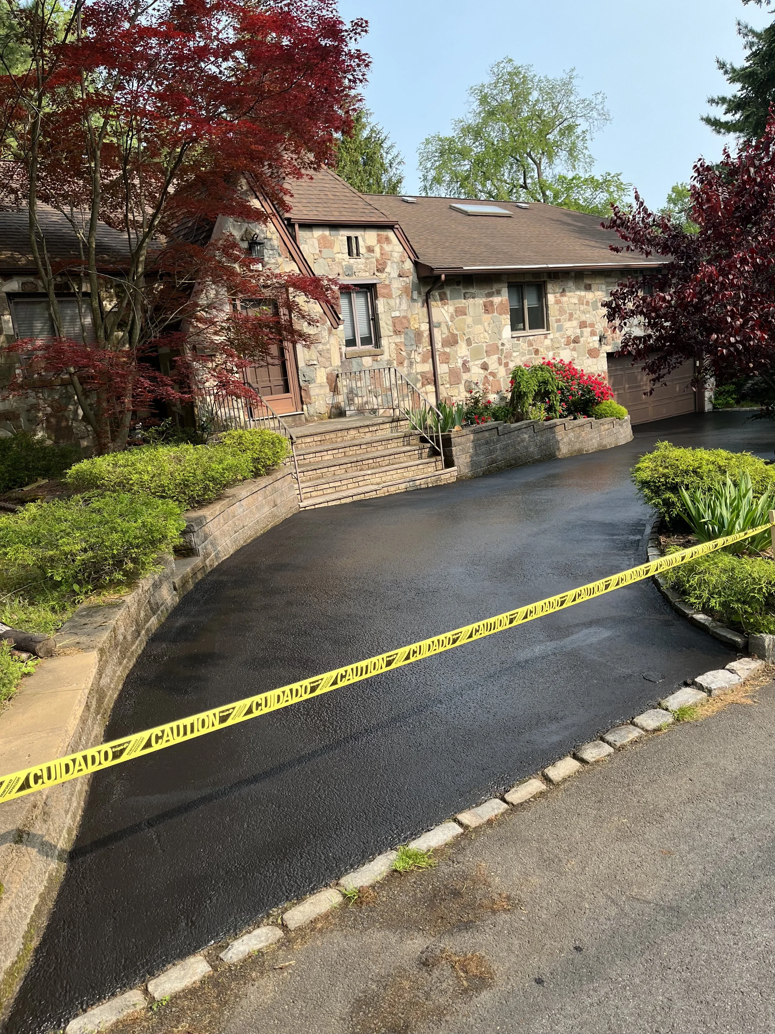 A newly paved black asphalt driveway leading to a house with stone exterior and a garage. Yellow caution tape is across the driveway indicating a possible hazard or caution area. The house has a front door, windows, and steps with a railing. Surrounding the driveway are landscaped plants and trees, some with red and green leaves. The sky is clear and blue.