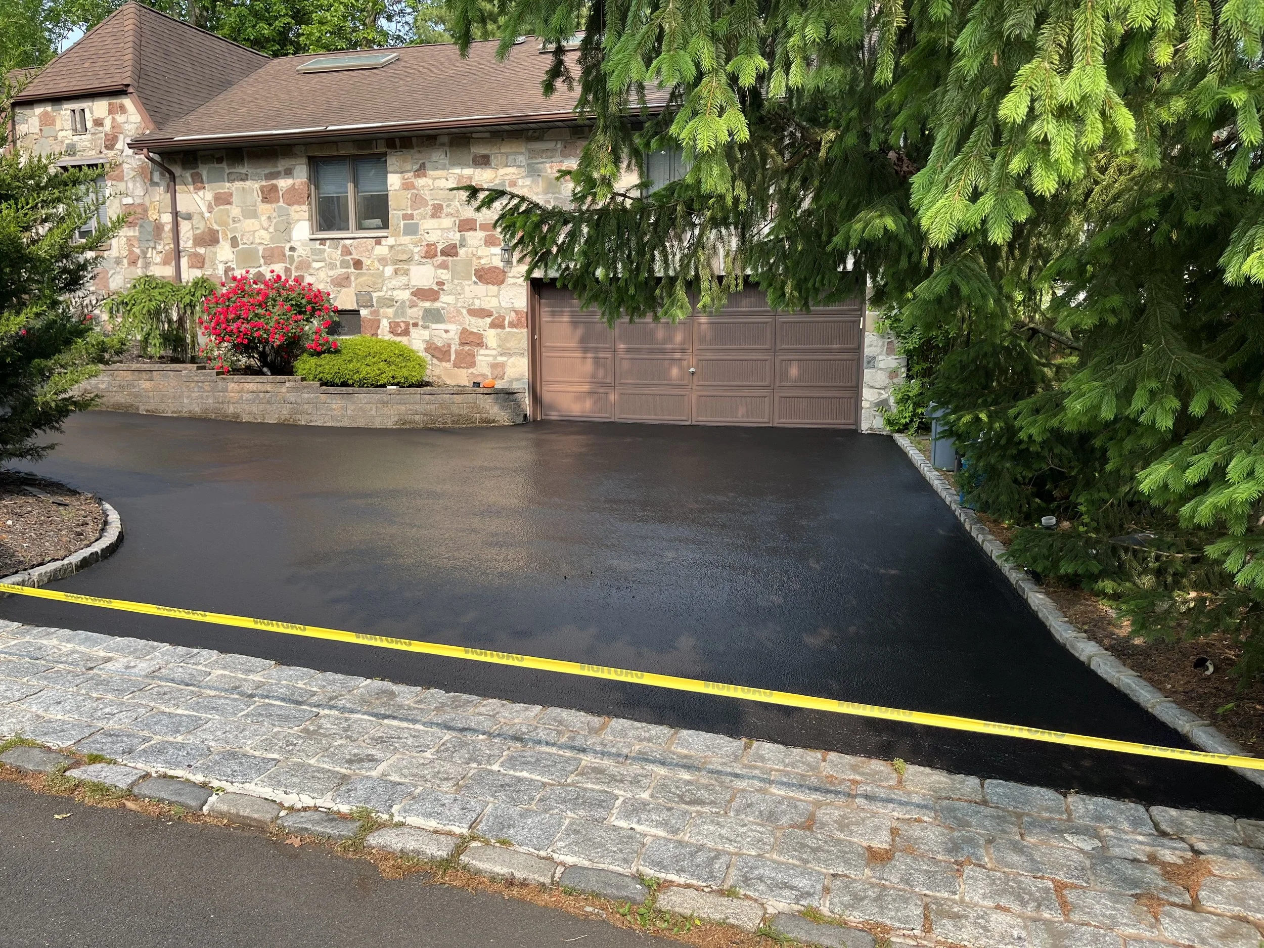 A residential house with a stone exterior, brown roof, and attached garage, partially obscured by large evergreen trees and a flower bed with pink and green plants. A yellow caution tape is laid out across the front of the paved driveway.