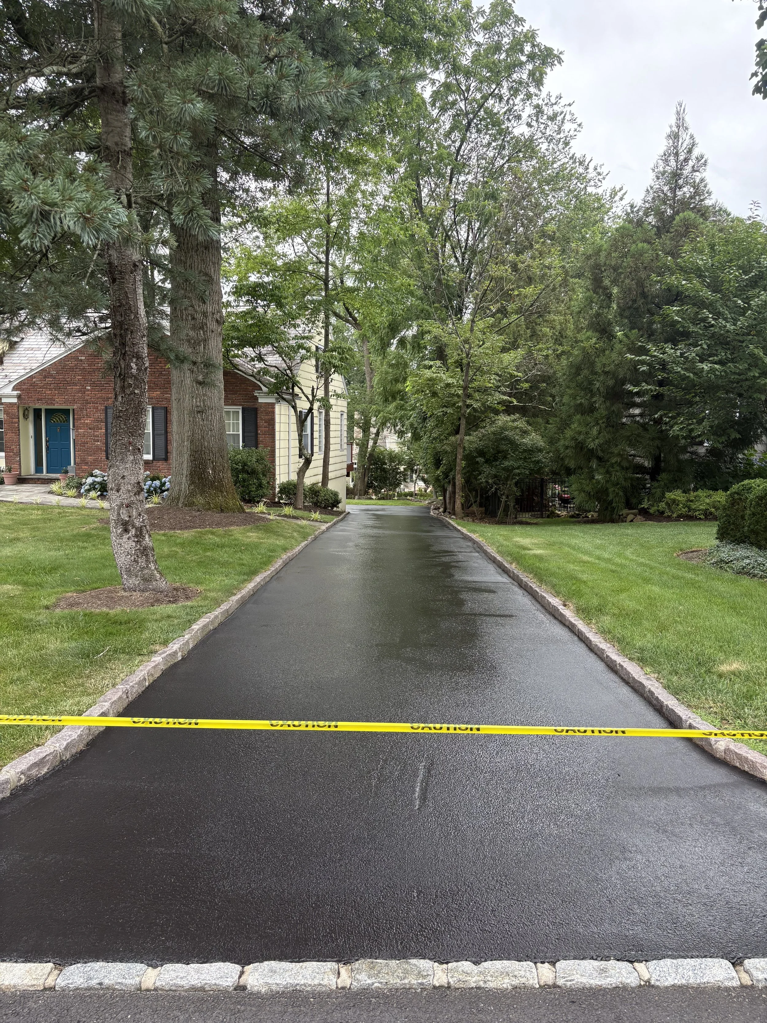 A freshly paved black asphalt driveway with yellow caution tape across it in a residential neighborhood with trees and houses, and a grassy lawn on both sides.