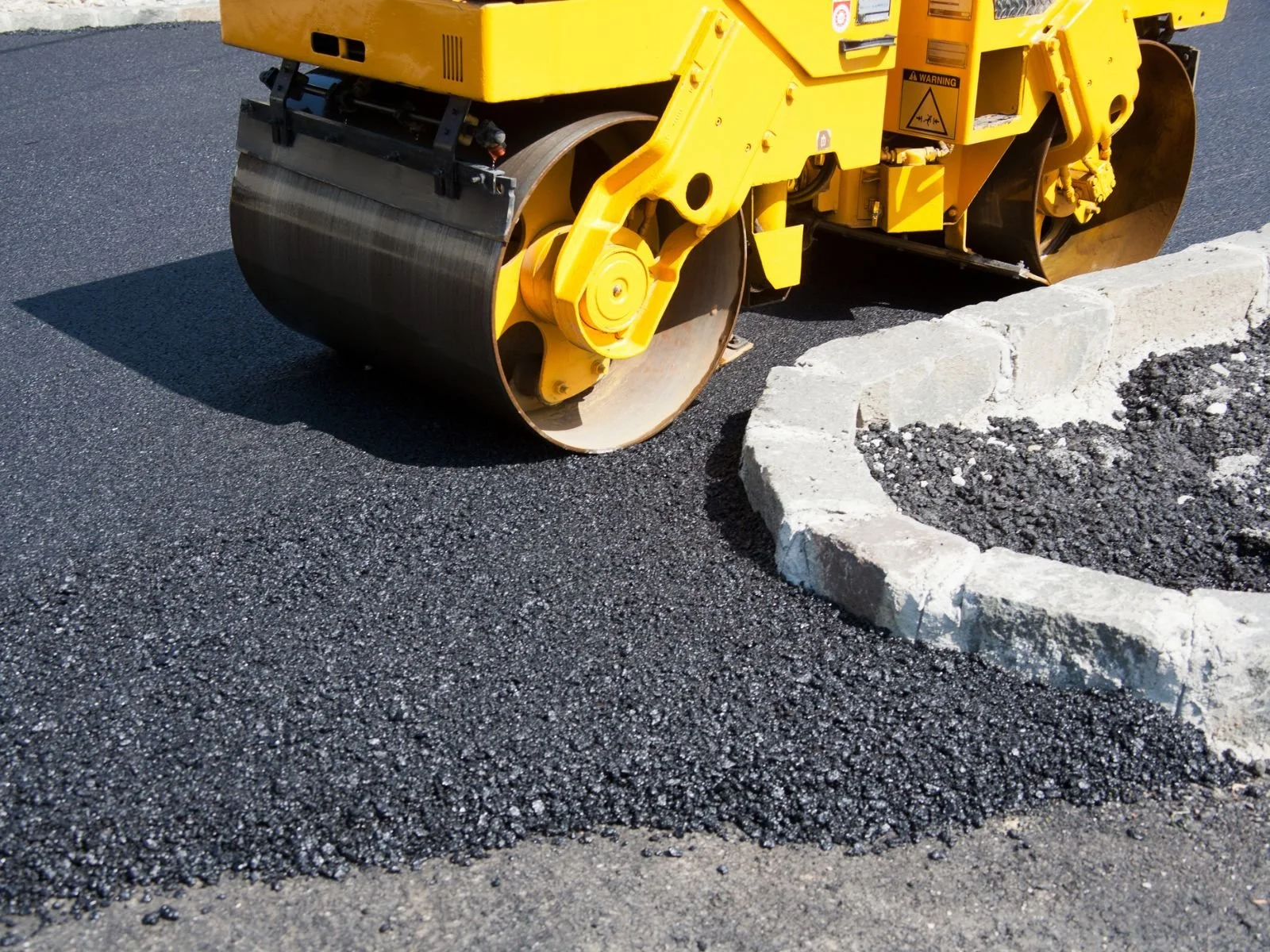 Close-up of a yellow road roller compacting asphalt on a road, with a section of a white concrete curb visible.
