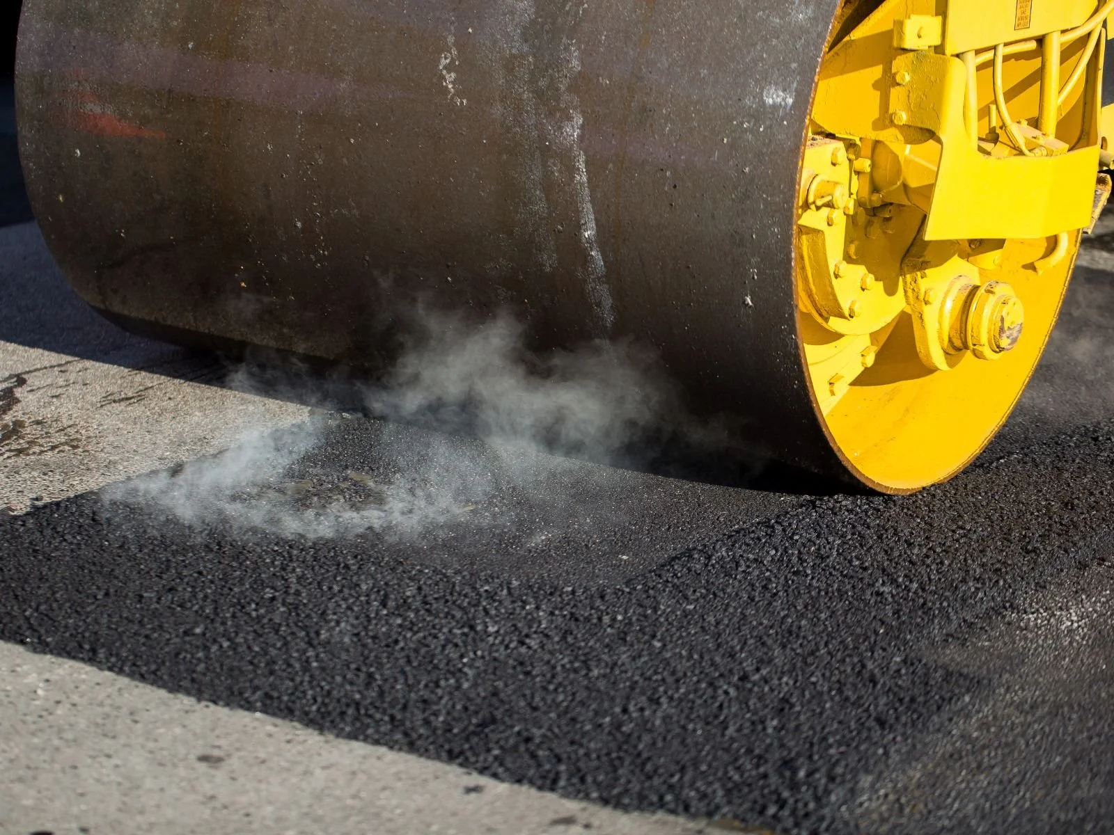 Close-up of a yellow road roller compacting asphalt on a road, with dust and smoke visible near the roller.