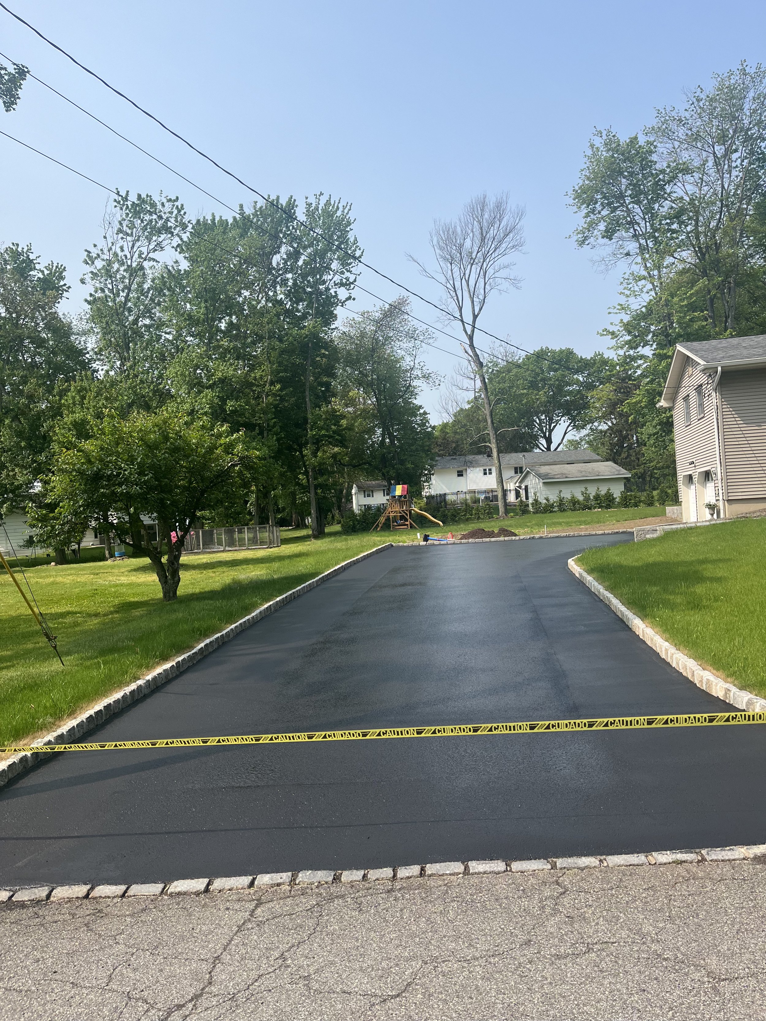 Freshly paved black asphalt driveway with yellow caution tape across the front, in a suburban neighborhood with houses, trees, and green lawns.