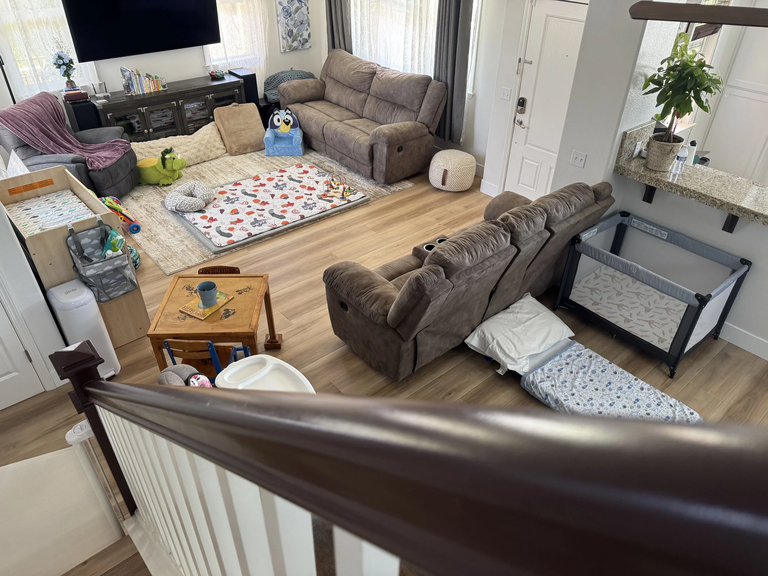 Living room with brown couches, a playpen, a crib with bedding, a small table with a plate and a mug, toys, a large TV, books, and a window with curtains.