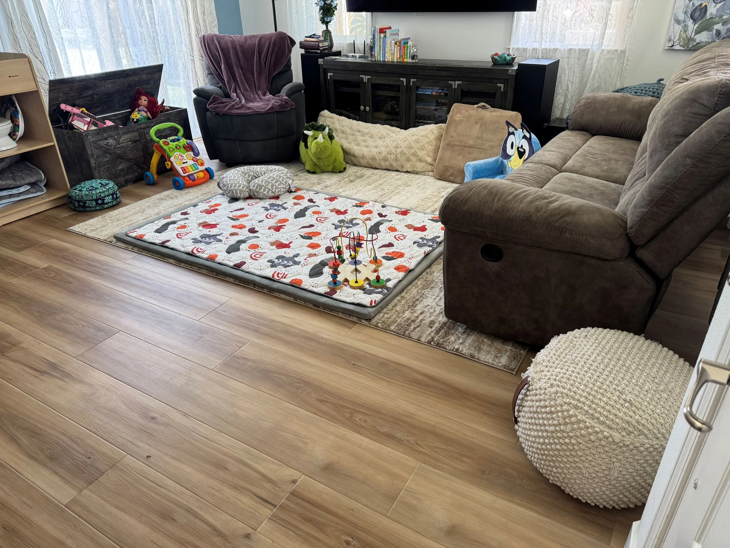 Living room with wooden floor, sofa, armchair with purple blanket, children's toys on a playmat, books on a shelf, and decorative pillows.