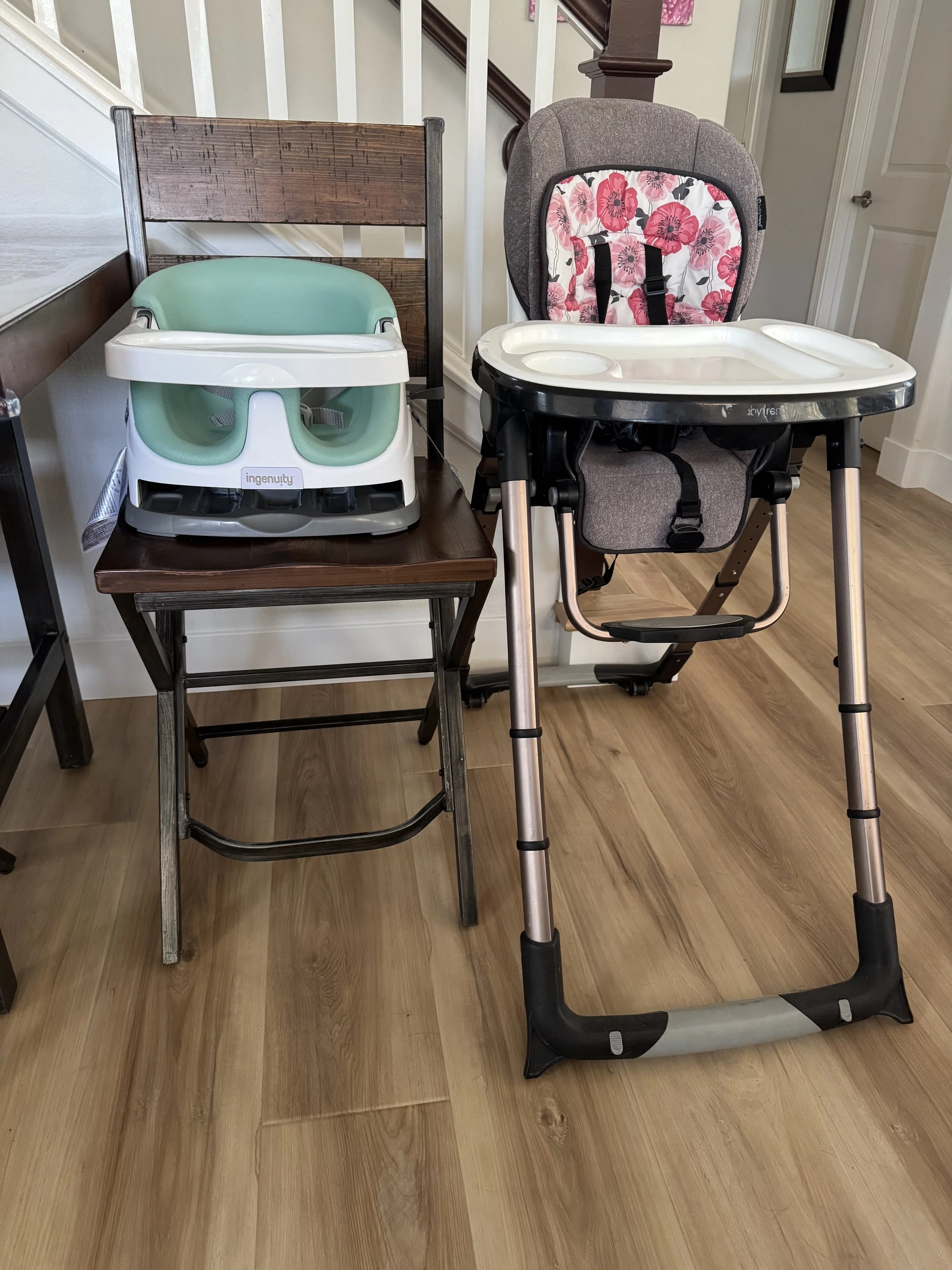 A wooden chair with a green and white baby bath seat on it and a dark gray high chair with pink floral pattern on its backrest in front of a staircase.