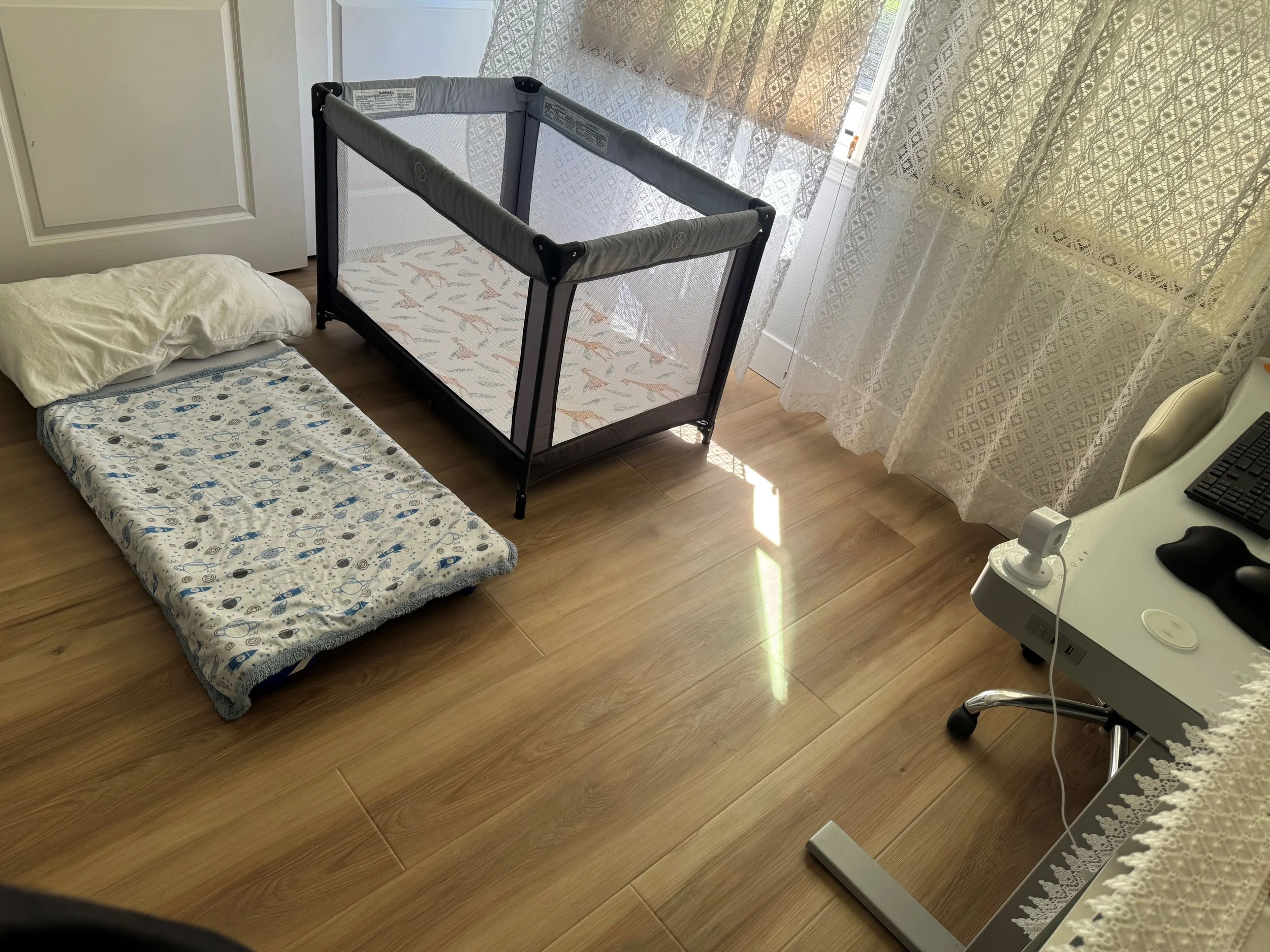A room with a wooden floor, a white door, a pillow and a blanket on the floor, a black and gray pet playpen, a small window with lace curtains, a white table with a computer and a mouse, and a white chair.