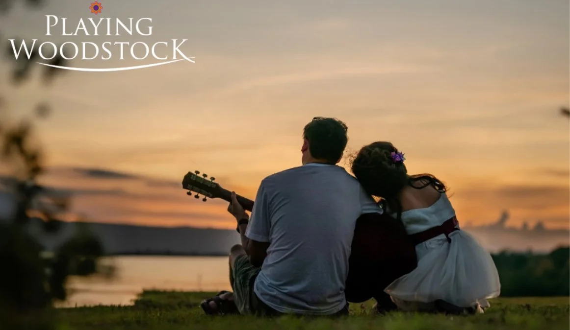A man and a young girl sitting together on grass by a lake at sunset, with the man playing guitar and the girl resting her head on his shoulder. Text reads "Playing Woodstock" with a small flower icon.
