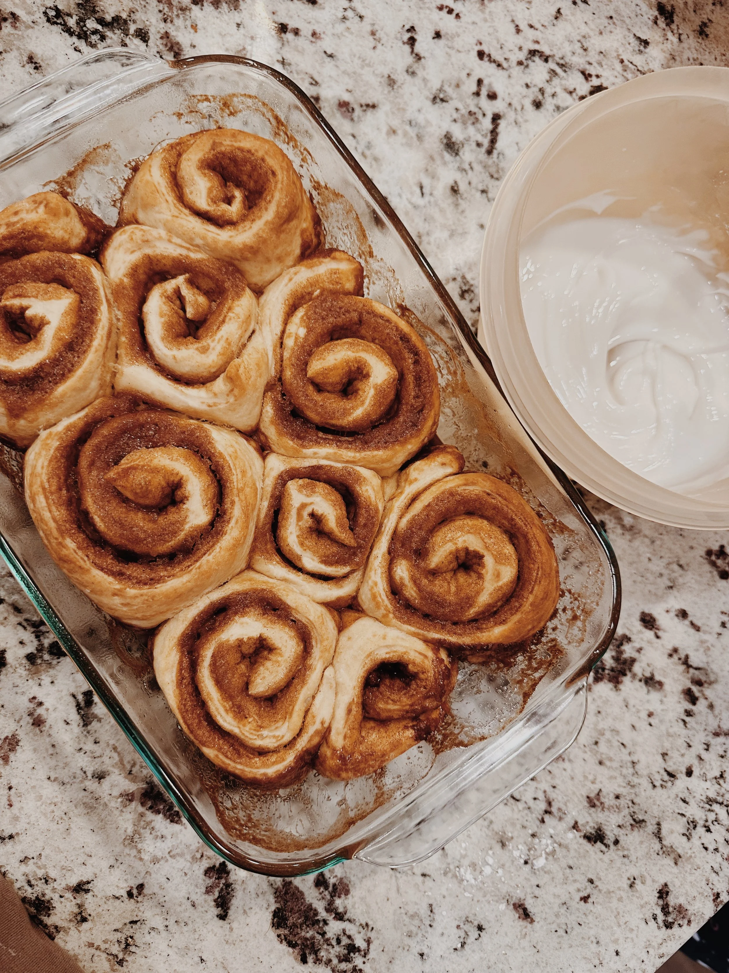 A glass baking dish filled with cinnamon rolls next to a container of cream cheese frosting on a speckled granite countertop.
