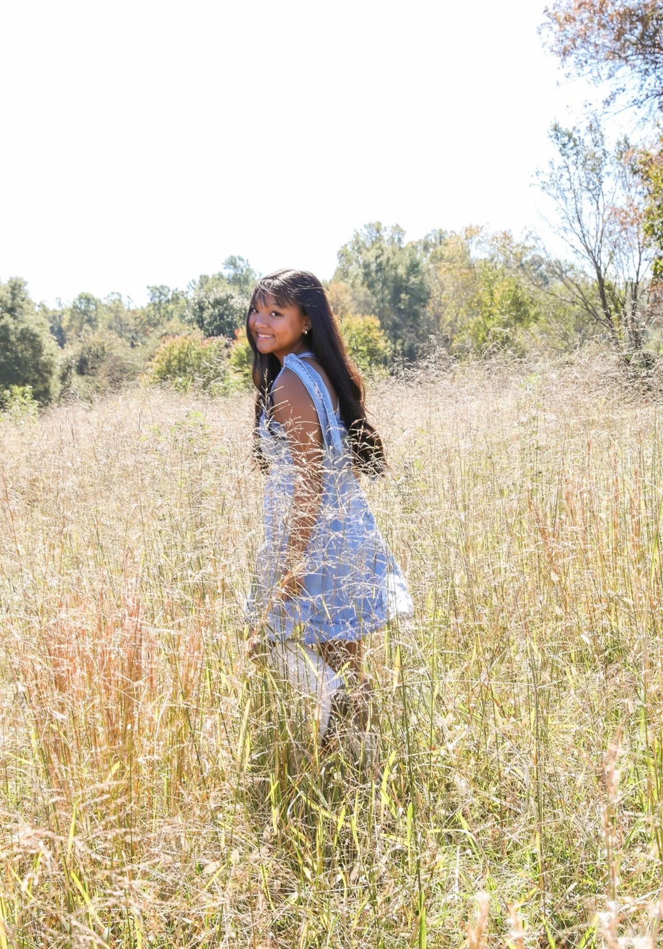Smiling young woman with long black hair wearing a denim dress standing in a field of tall grass with trees in the background on a sunny day.