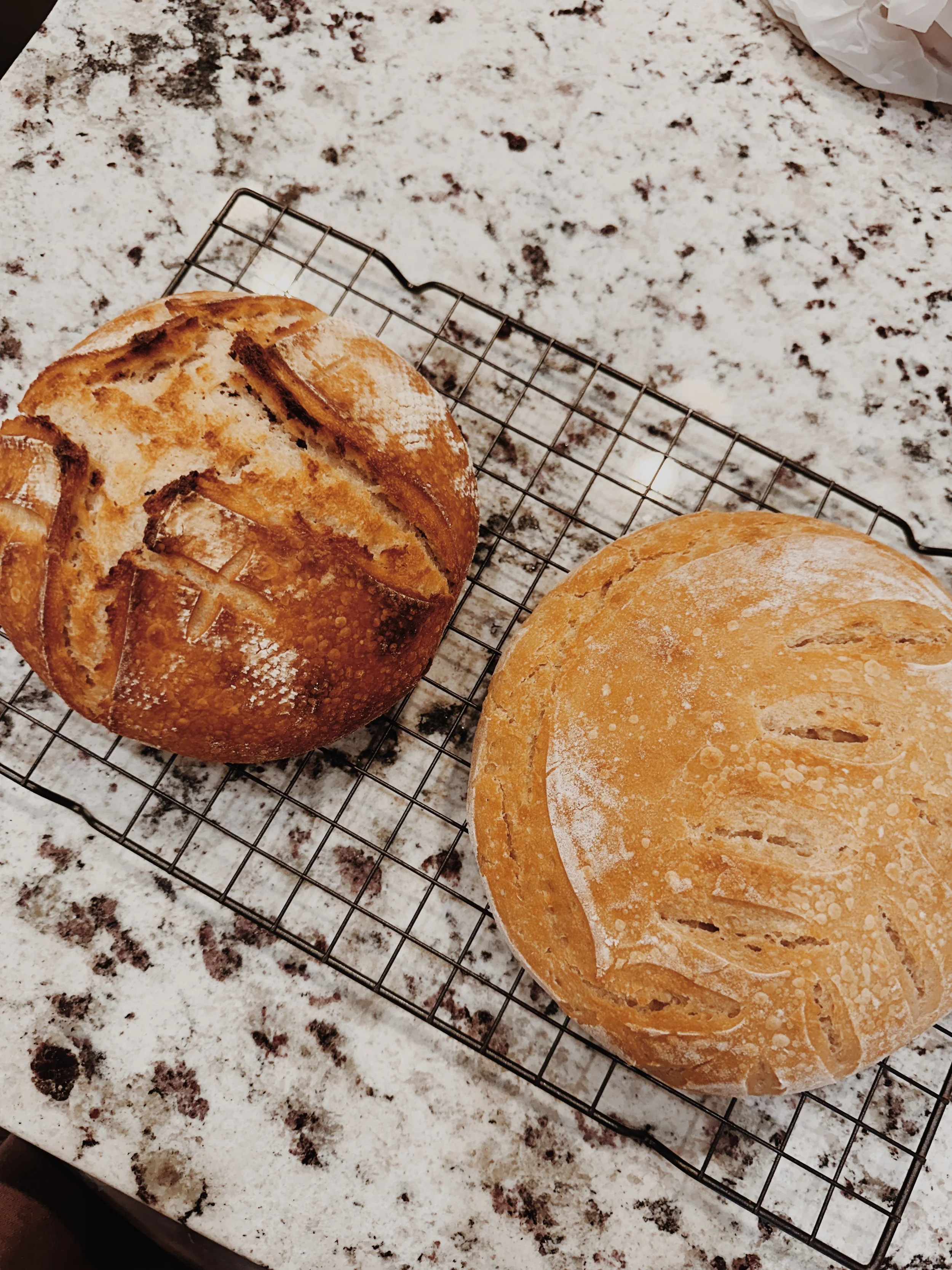 Two loaves of bread cooling on a wire rack on a speckled granite countertop, one with a round shape and decorative scoring, the other slightly flattened with a simple scoring pattern.