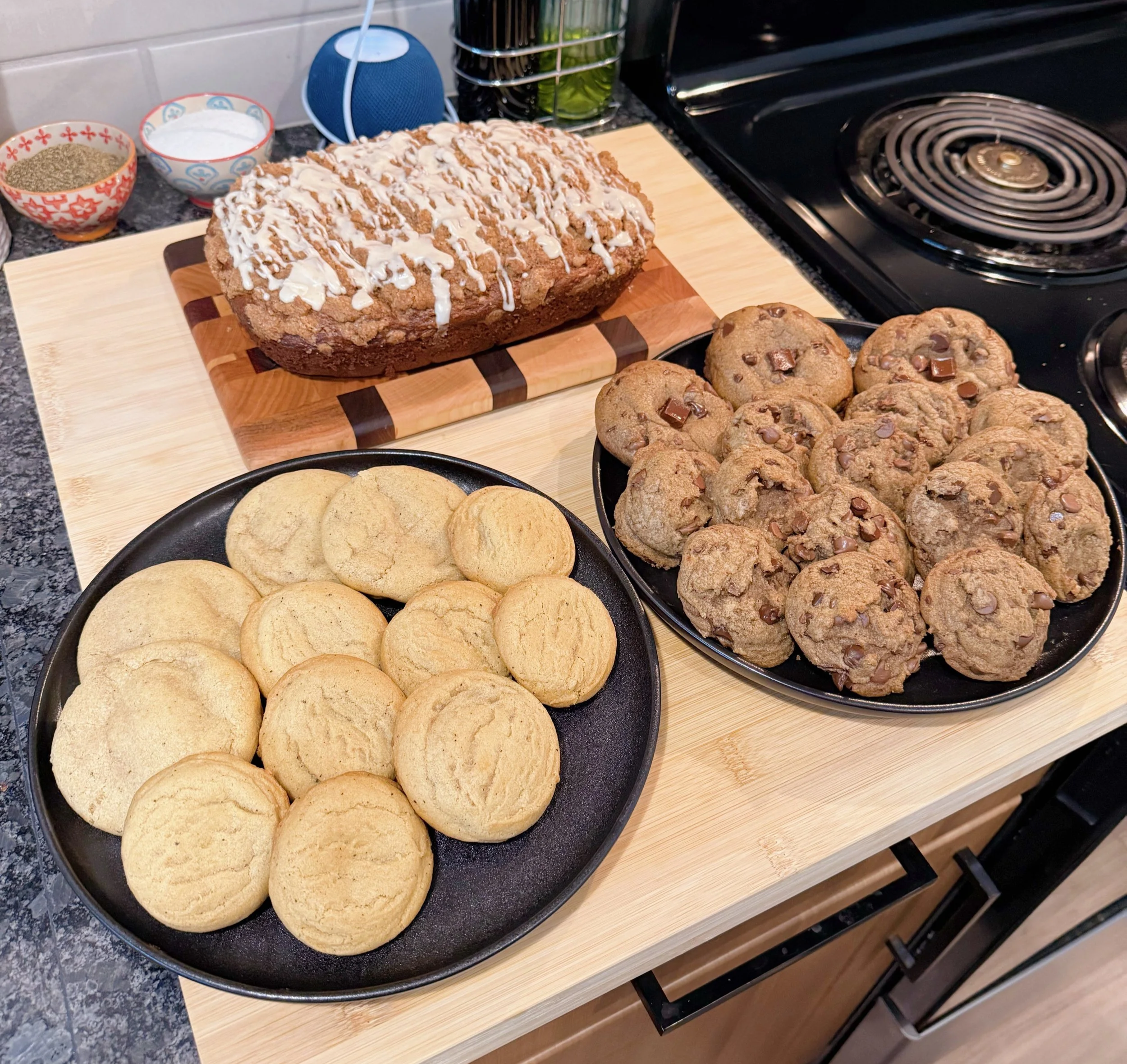 Assorted cookies and baked goods on a kitchen countertop, including a plate of sugar cookies, a plate of chocolate chip cookies, and a loaf of bread with icing drizzle.