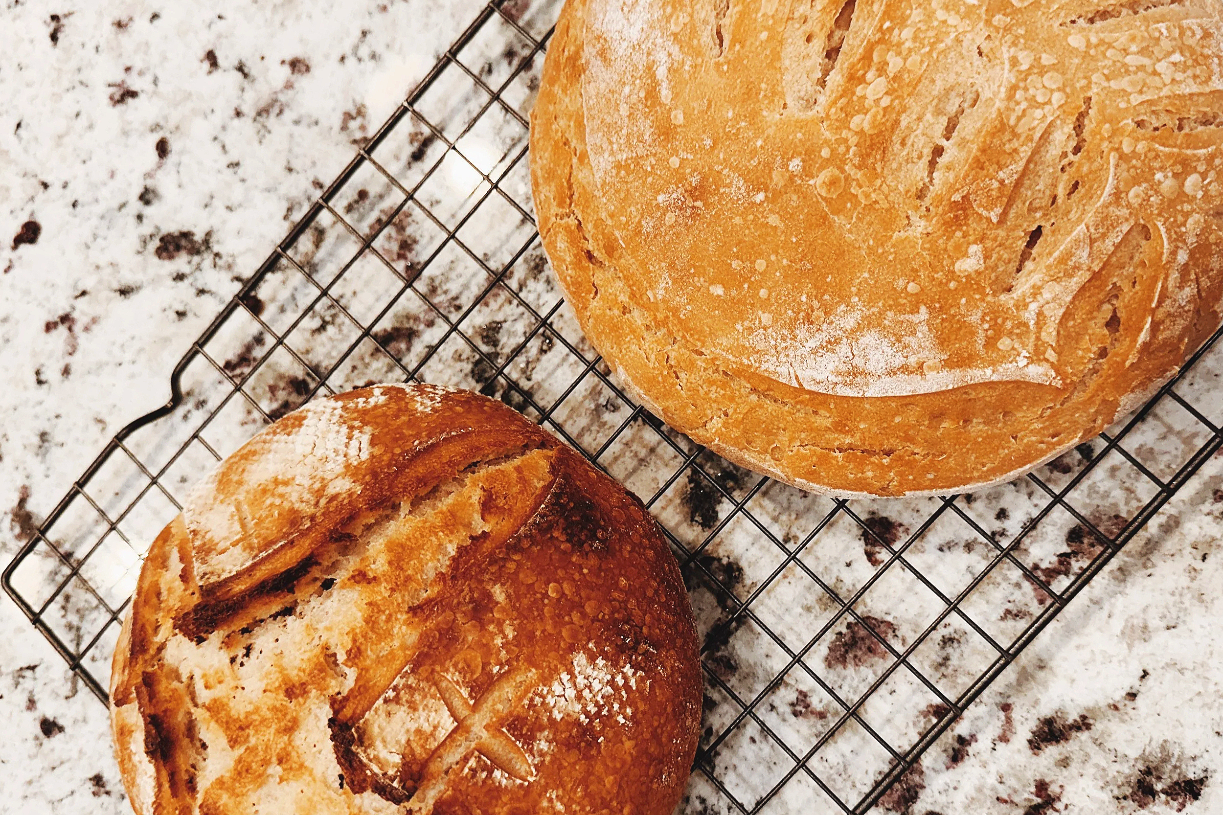 Two freshly baked loaves of bread on a wire cooling rack on a marble countertop.
