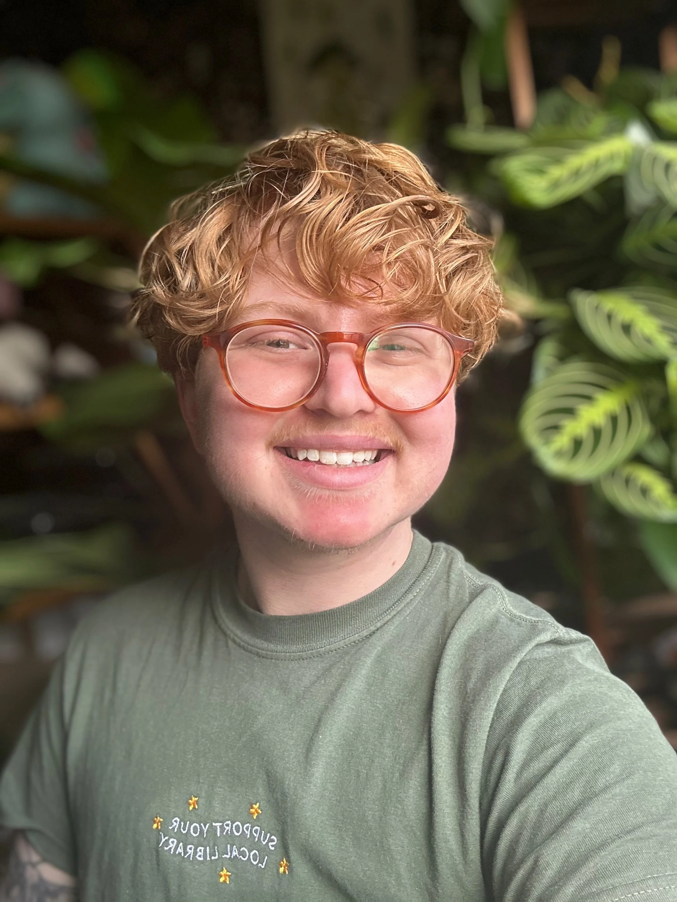 Close-up of a young person with curly orange hair, wearing glasses, smiling, and wearing a green shirt with text and stars, standing in front of green leafy plants.