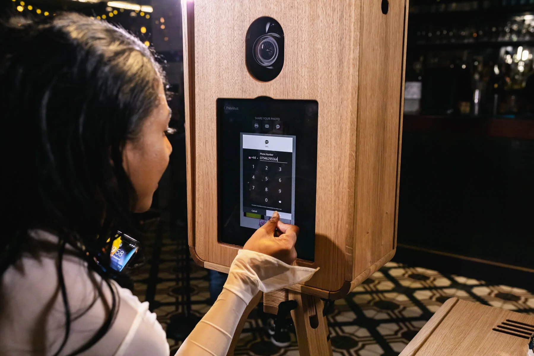 A woman is entering a phone number on an electronic touchscreen device mounted on a wooden stand, with a camera above the screen, in an indoor setting with dim lighting.