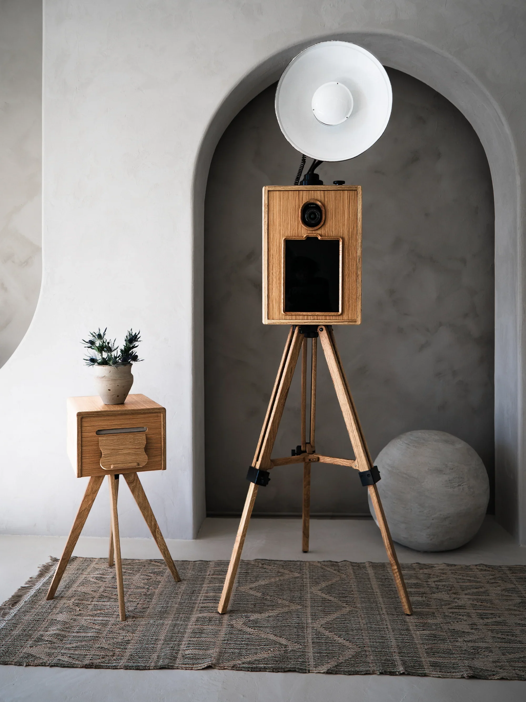 A photography studio setup with a wooden camera on a tripod, a large studio light with a white reflector, a small wooden side table with a potted plant, a rug on the floor, and a large spherical object in the background next to a textured curved wall.