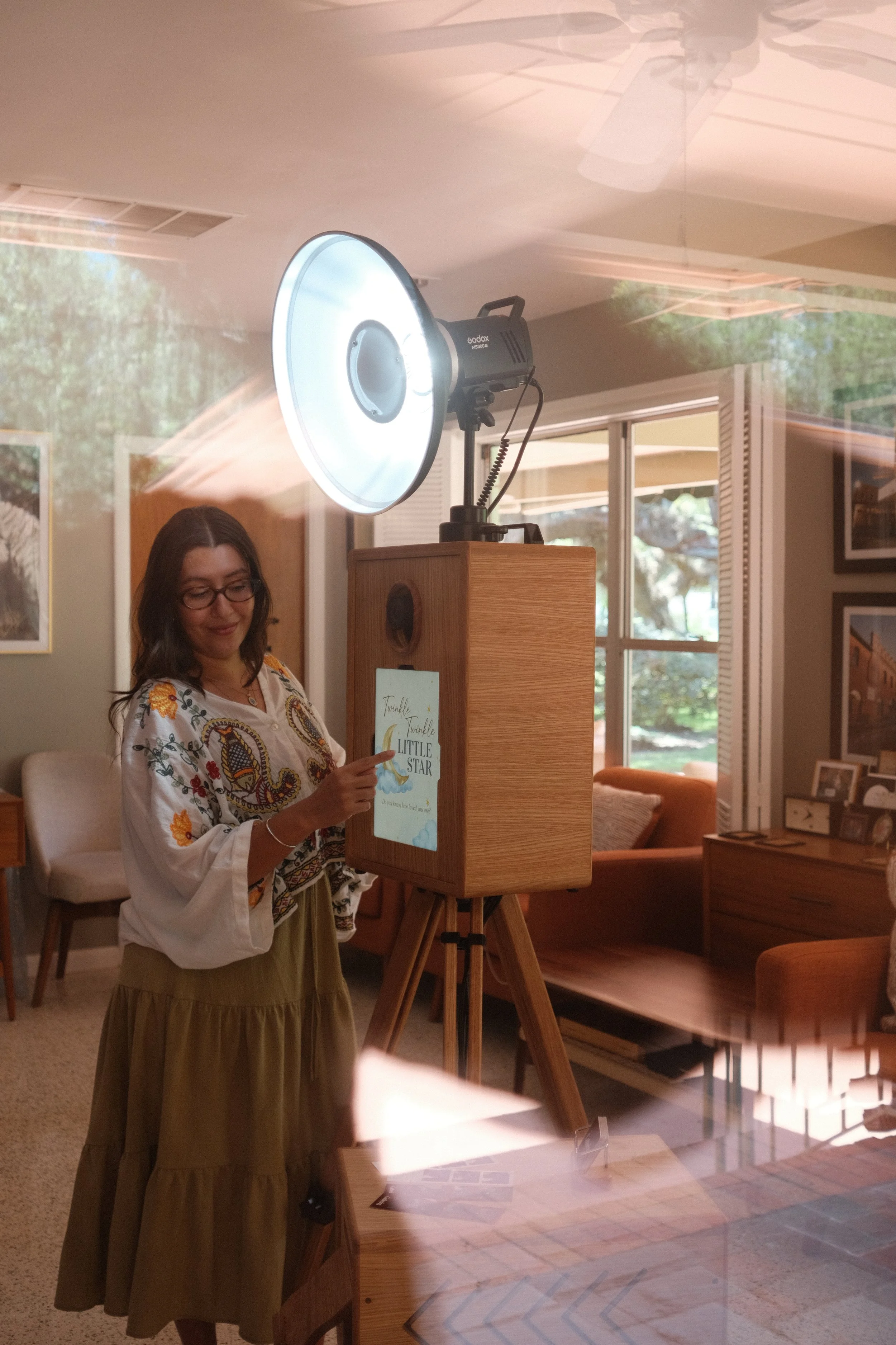 A woman with glasses and a floral top is looking at a book titled 'Twinkle Twinkle Little Star' displayed on a wooden stand inside a cozy room with large windows, trees visible outside, framed photos on the wall, and a ceiling fan above.