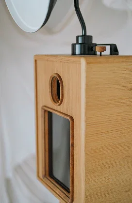 Wooden speaker with a black power cord plugged into the top, placed near a mirror on a white wall.