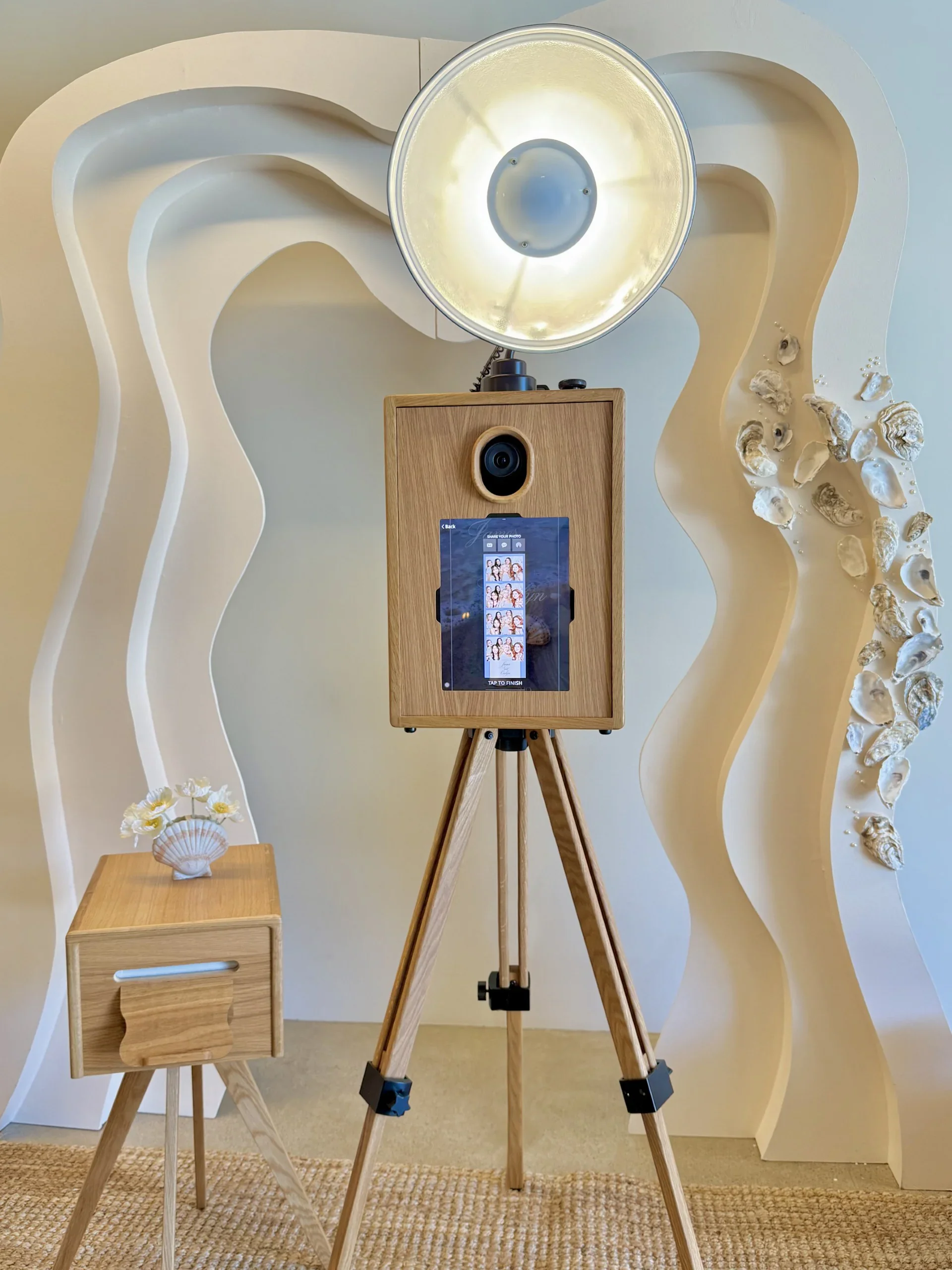 A photo booth setup with a wooden tripod stand, a wooden table with a shell decoration, a mirror, a photo strip on a screen, and decorative shells on a wall in the background.