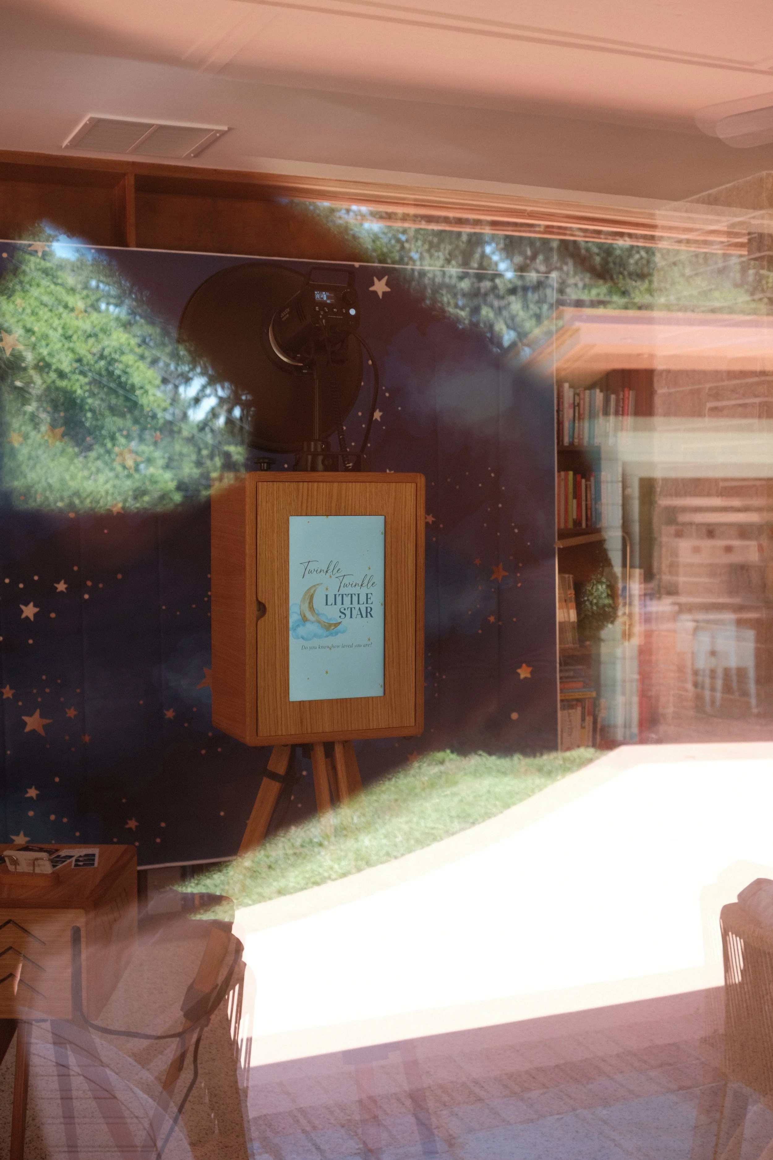 Indoor scene with a wooden easel holding a framed sign that reads "Twinkle Twinkle Little Star" and a backdrop decorated with stars and a moon, with bookshelves and a window showing trees outside.