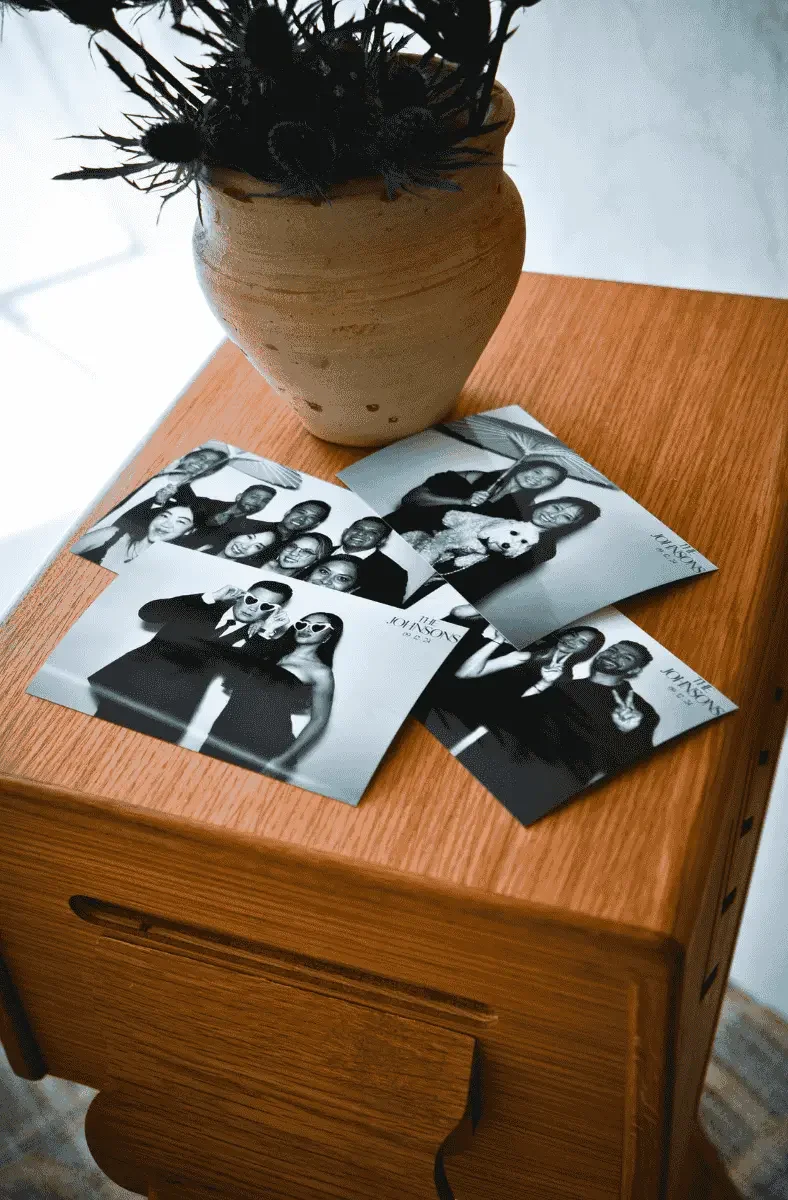 Black and white photo booth pictures, some with people wearing sunglasses and posing with friends, resting on a wooden table next to a clay vase with black feathers.