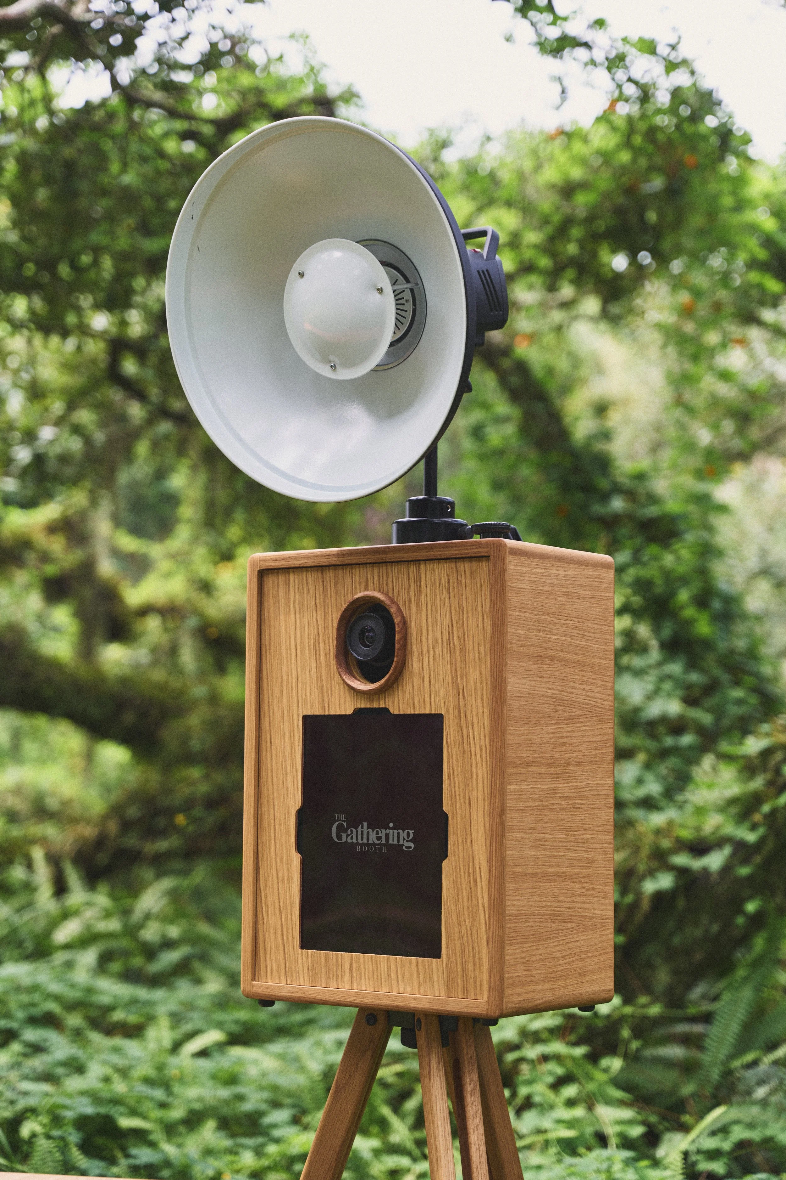 A wooden photo booth with a black chalkboard area, labeled "The Gathering Booth," set outdoors amidst green foliage.