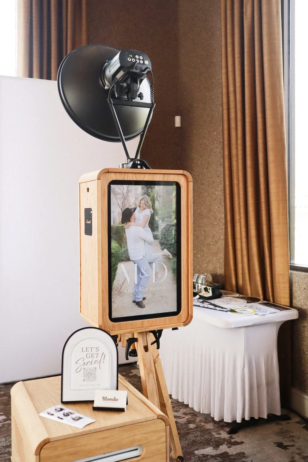 Photo booth with a wooden frame and a screen displaying a couple hugging, advertising a wedding of Macey and Dakota, with a camera on top and decorative signage nearby.