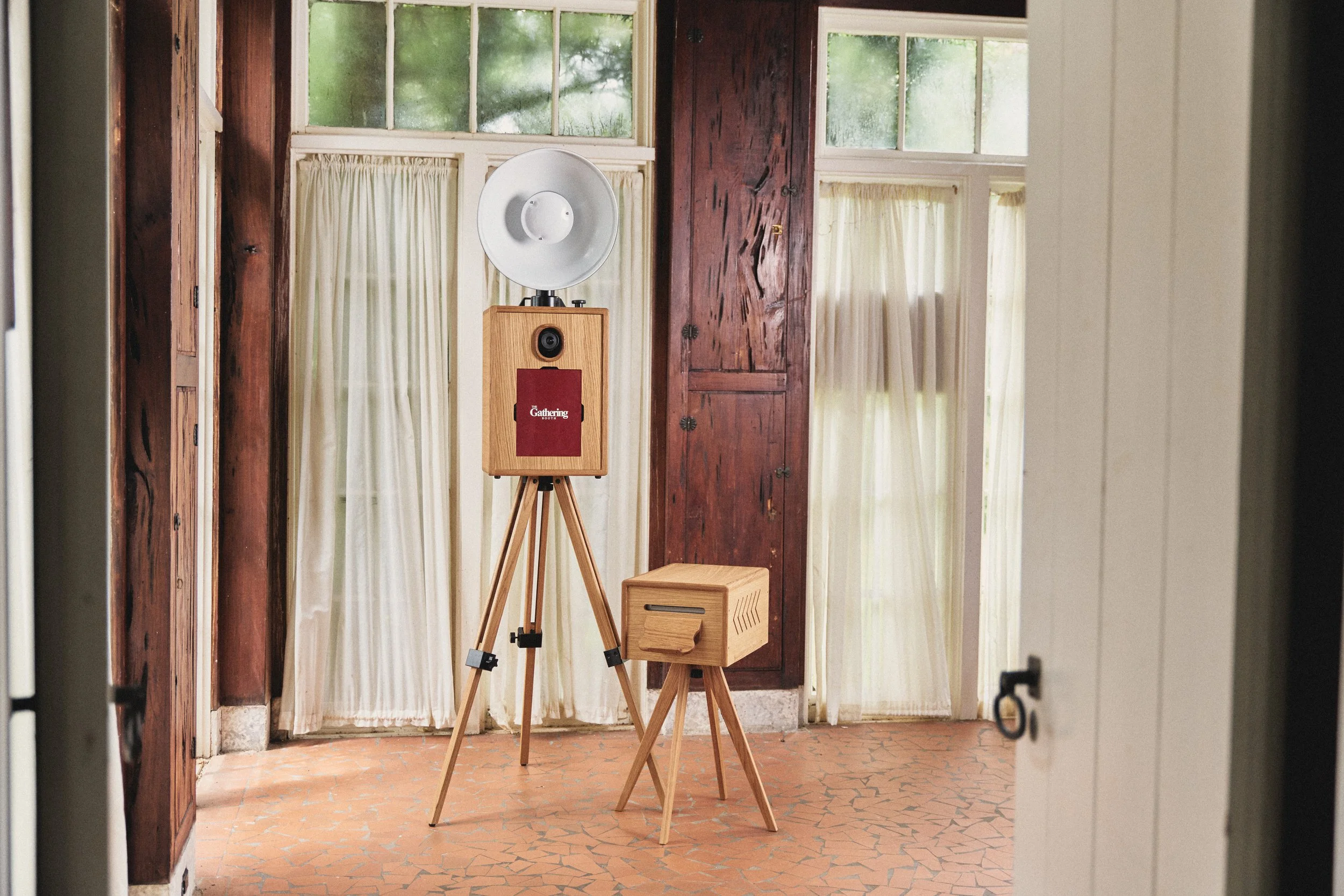 An interior view of a room seen through a doorway, with a wooden speaker on a tripod, a smaller wooden stool, and an open wooden cabinet with a window in the background showing greenery outside.