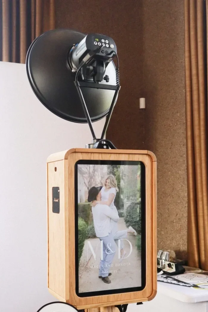A wooden photo booth with a digital screen displaying a couple embracing, with the text 'M & D' and 'Lacey and Dakota' at the bottom, and a black professional studio light mounted above.