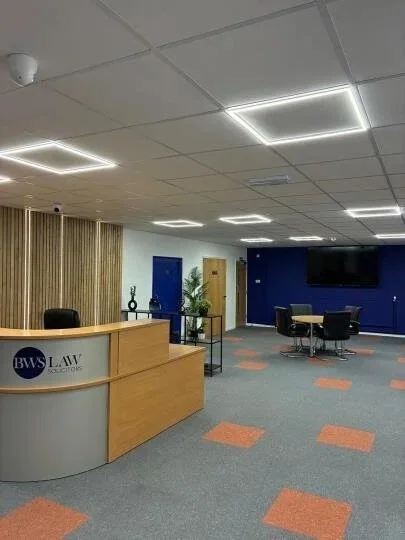 Office reception area with a wooden desk displaying a BWS Law Solutions logo, a small table with decorative items, a blue and yellow door, a small table with chairs, a wall-mounted TV, and a blue accent wall.