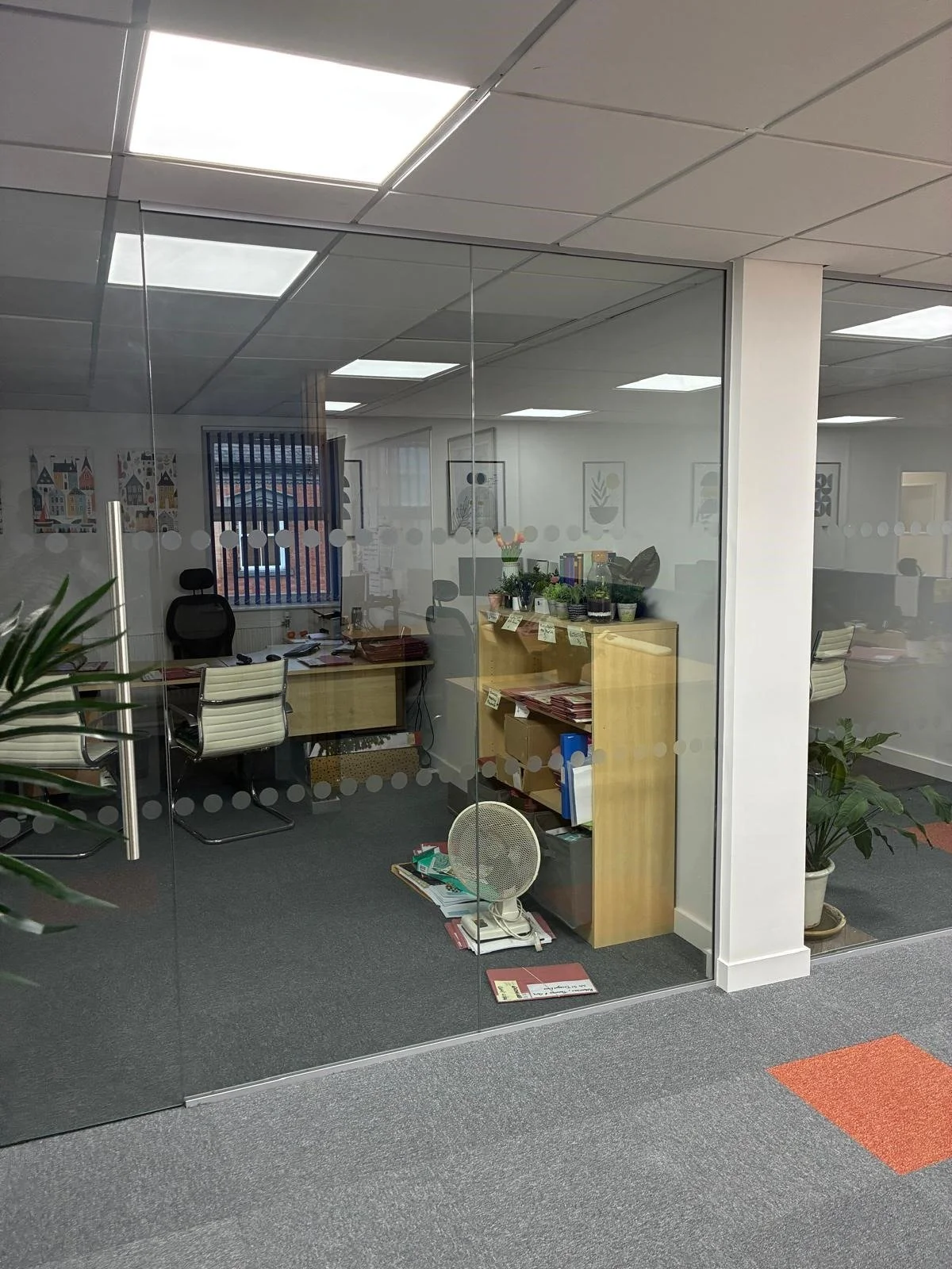 View of an office space through glass walls, showing desks, chairs, potted plants, and office supplies.