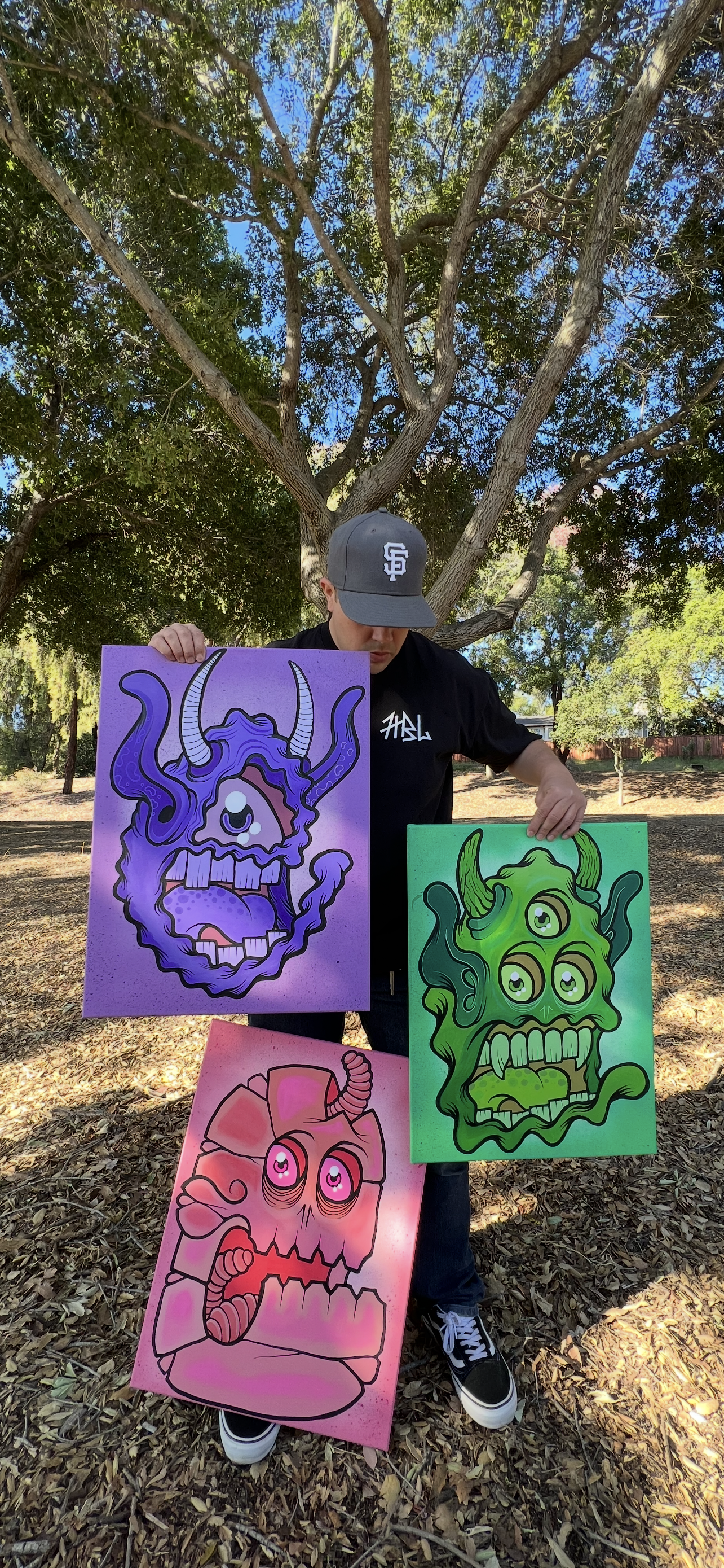 Person wearing a gray cap with a white logo, black shirt, and black and white sneakers standing outdoors under a large tree, holding three colorful monster-themed paintings with three eyes, horns, and exaggerated features, on a leaf-covered ground.