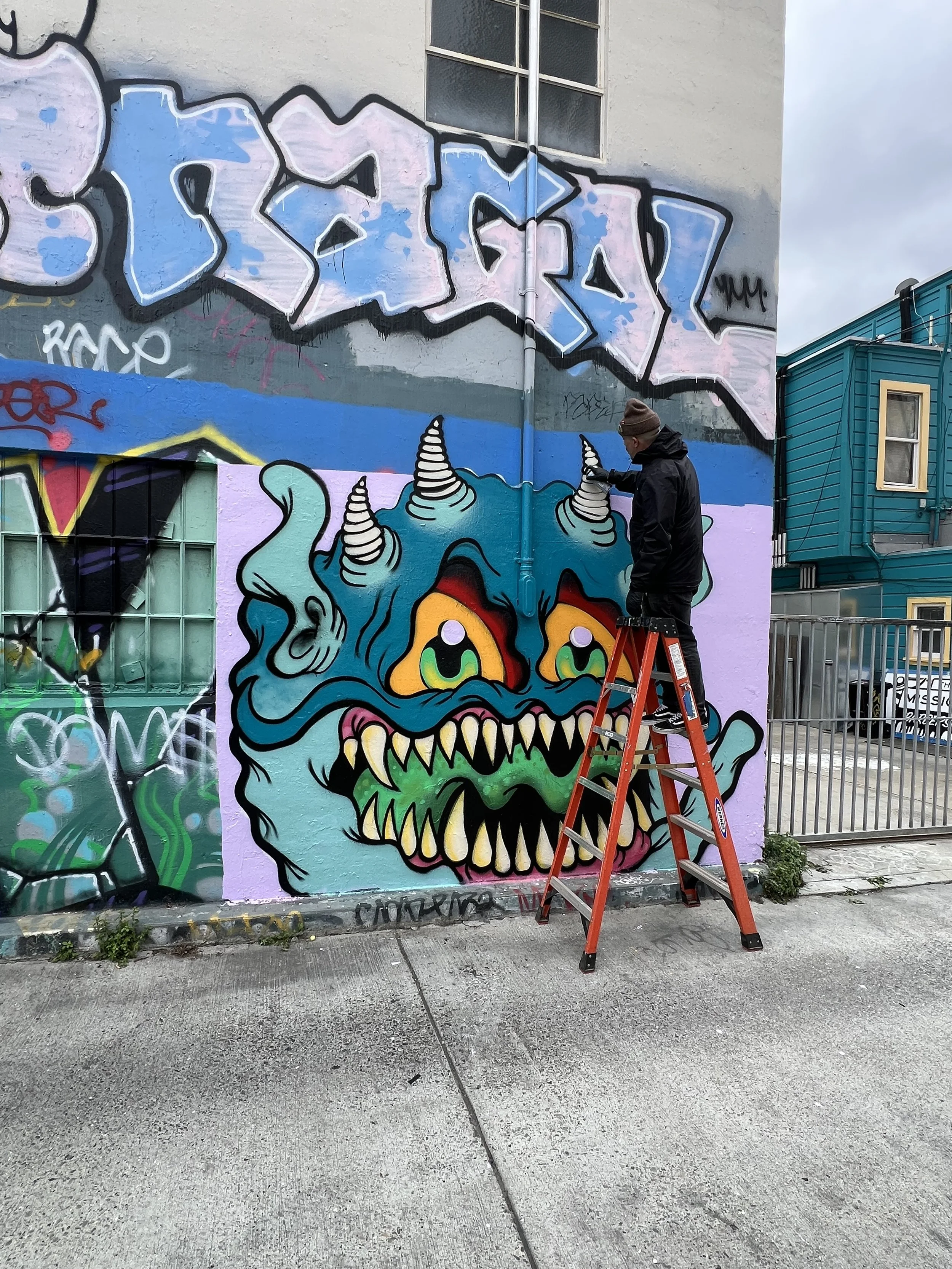 A person standing on an orange ladder painting a large colorful mural of a cartoonish monster face on a building wall. The monster has teal skin, sharp yellow teeth, and spiral horns. The wall features additional graffiti and street art.