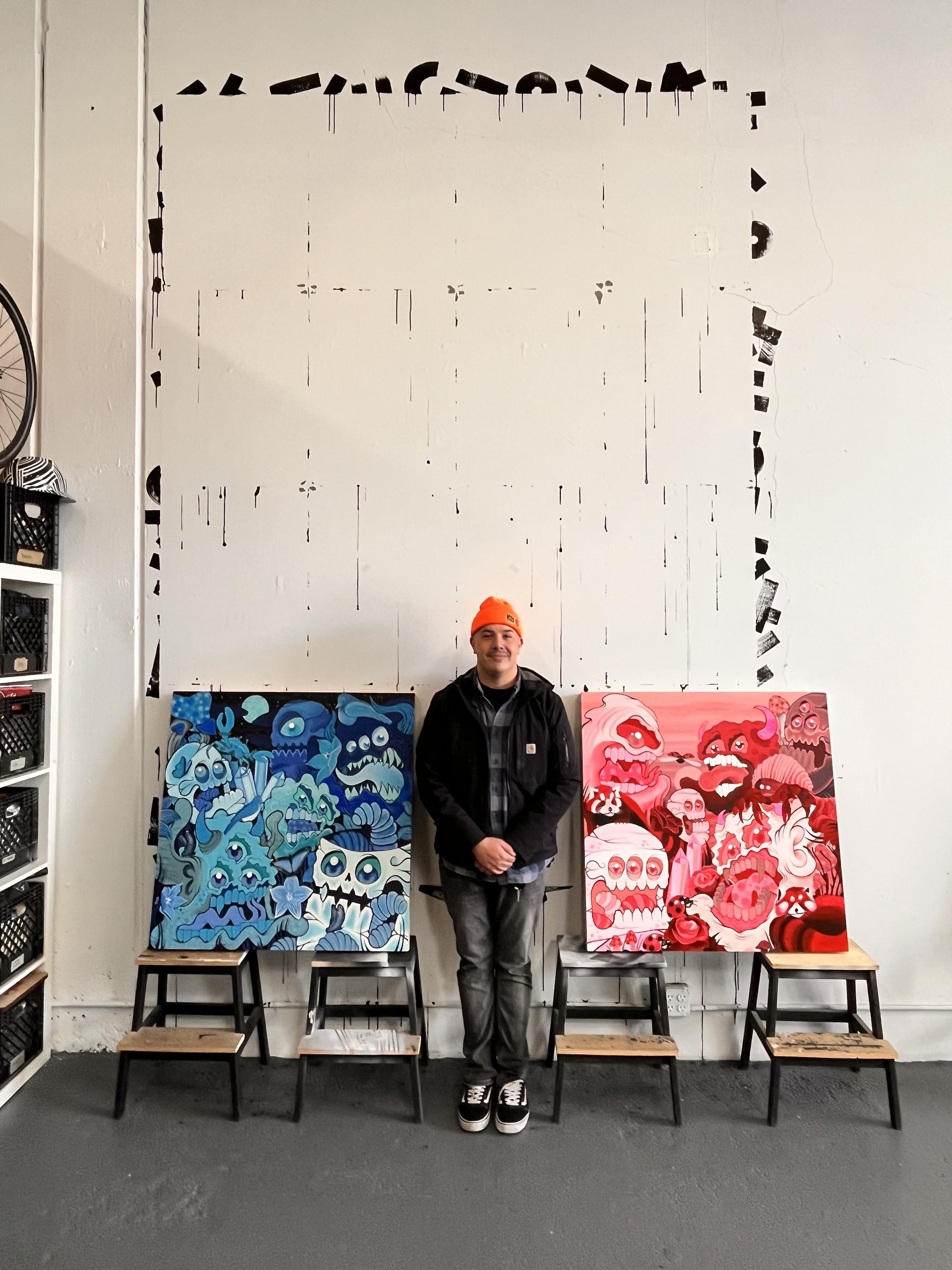 A person wearing a bright orange beanie hat and dark jacket standing in front of two colorful paintings of skulls with floral and abstract designs, inside an art gallery or studio with stools and a white wall.
